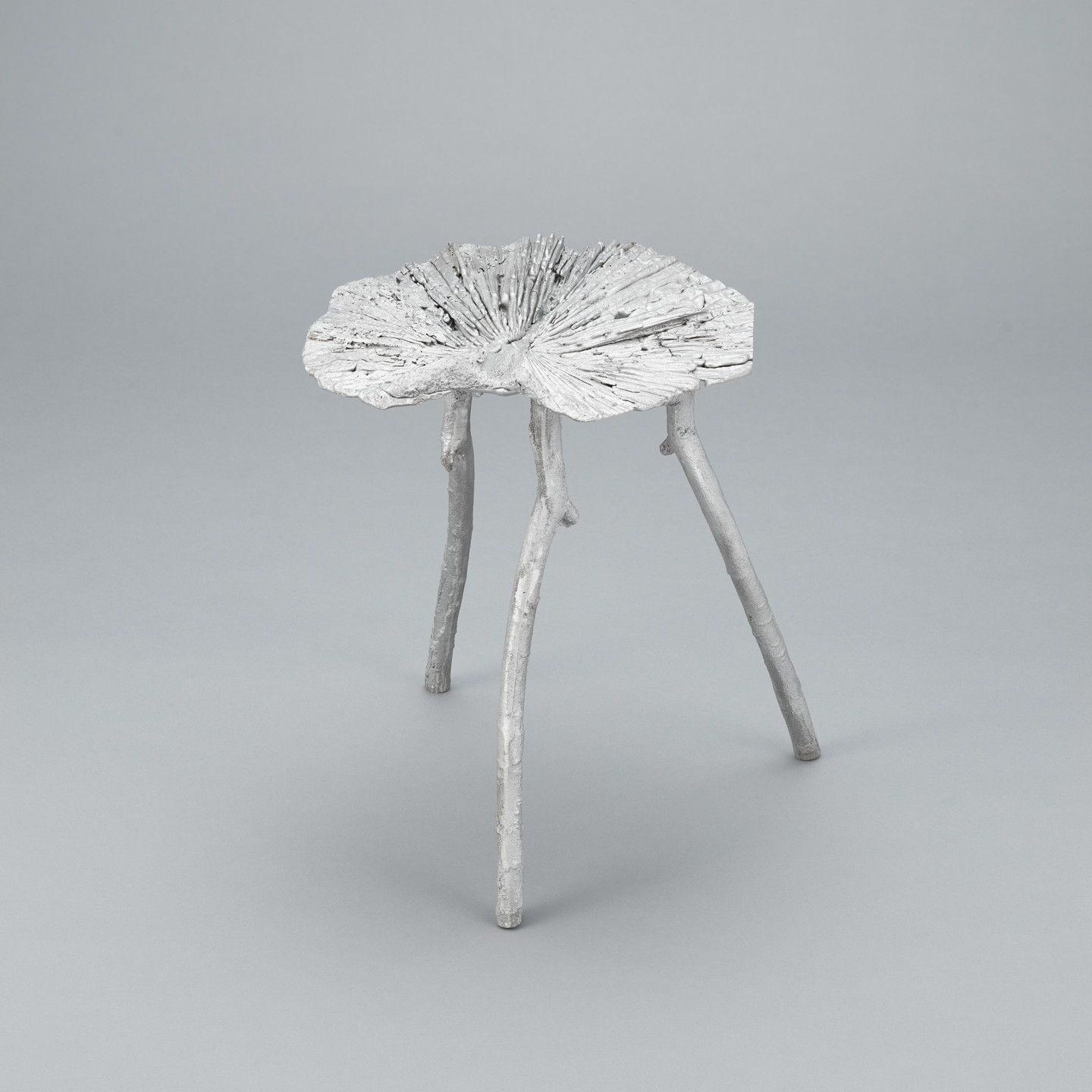 A three-legged stool is shaped like a large, textured leaf with branched legs, all in a light gray color. The background is a plain, lighter gray surface. This is an AI generated description. To give feedback, email ai@moma.org.