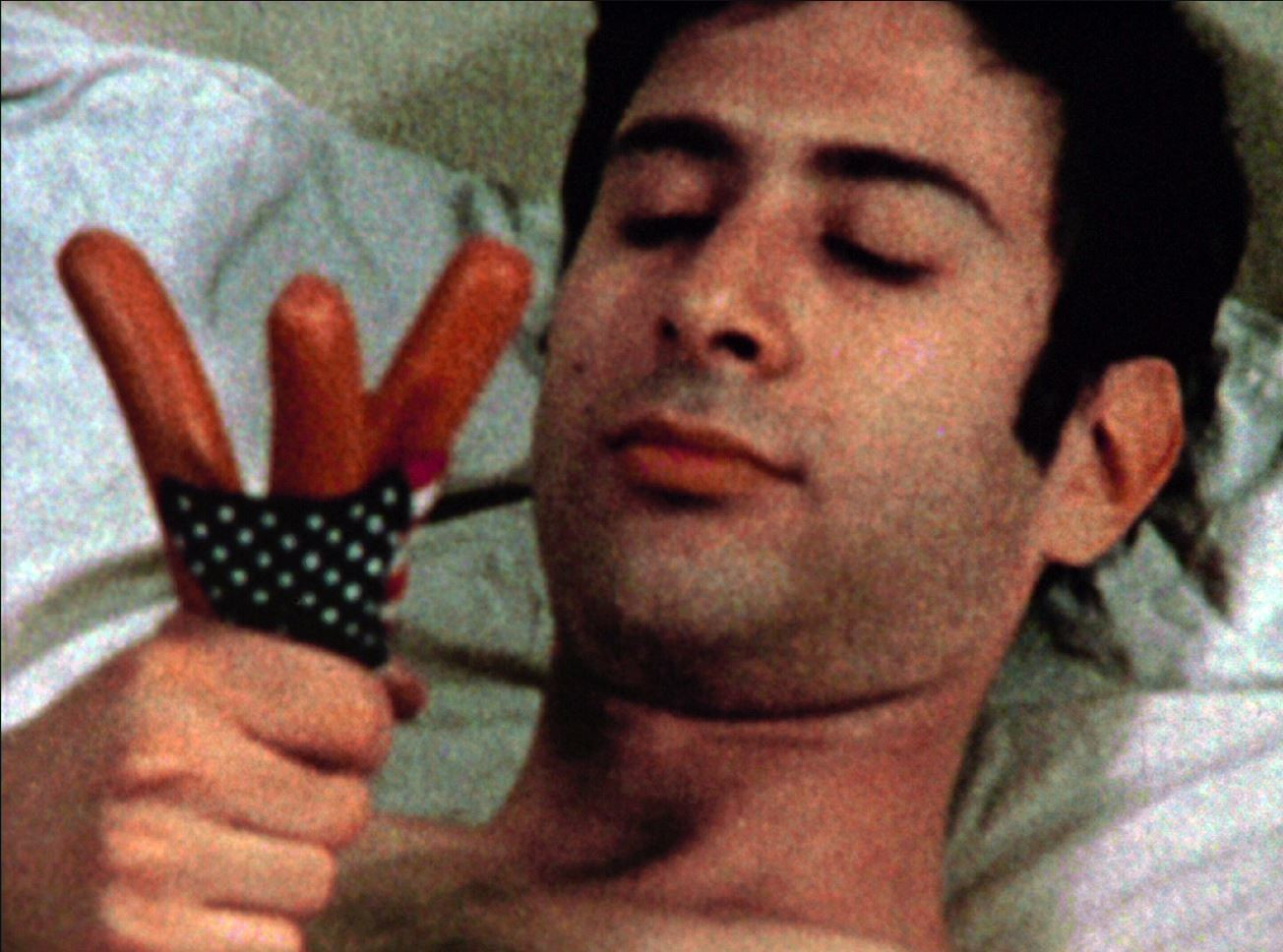 A person with closed eyes lies in bed, holding three hot dogs between their fingers, which have a polka-dotted cloth wrapped around them. The predominant colors in the image are shades of brown and white. This is an AI generated description. To give feedback, email ai@moma.org.