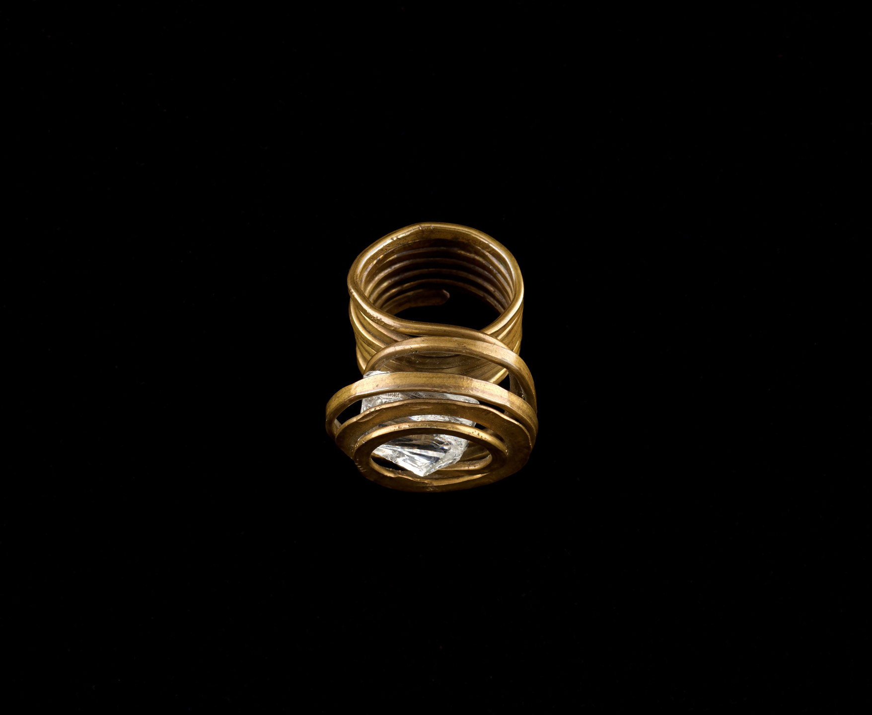 A gold ring with a wide, spiral band is seen against a black background. The ring features a large, clear gemstone set within the spiral. This is an AI generated description. To give feedback, email ai@moma.org.