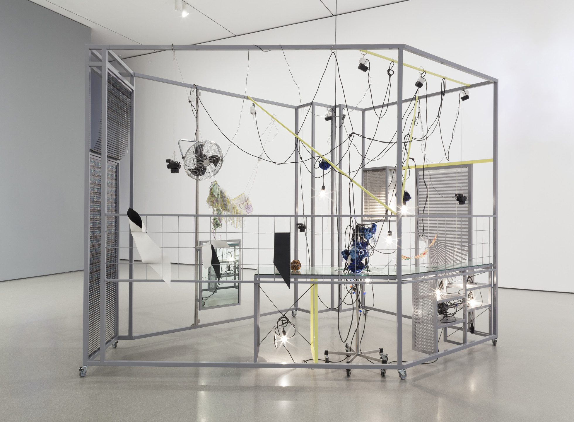 A grey metal frame structure filled with tangled black wires, light bulbs, various small fans, and hanging objects sits in a white room with a smooth floor. Some parts of the frame are painted yellow and a monitor displays a reflection within the structure. This is an AI generated description. To give feedback, email ai@moma.org.