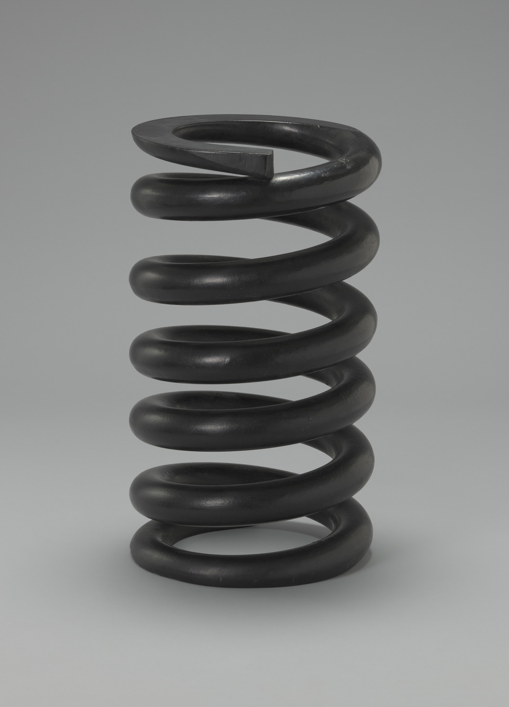 A black, coiled, metal spring with roughly five loops stands upright against a neutral gray background. The spring is thick and well-defined, with a smooth, glossy surface. This is an AI generated description. To give feedback, email ai@moma.org.