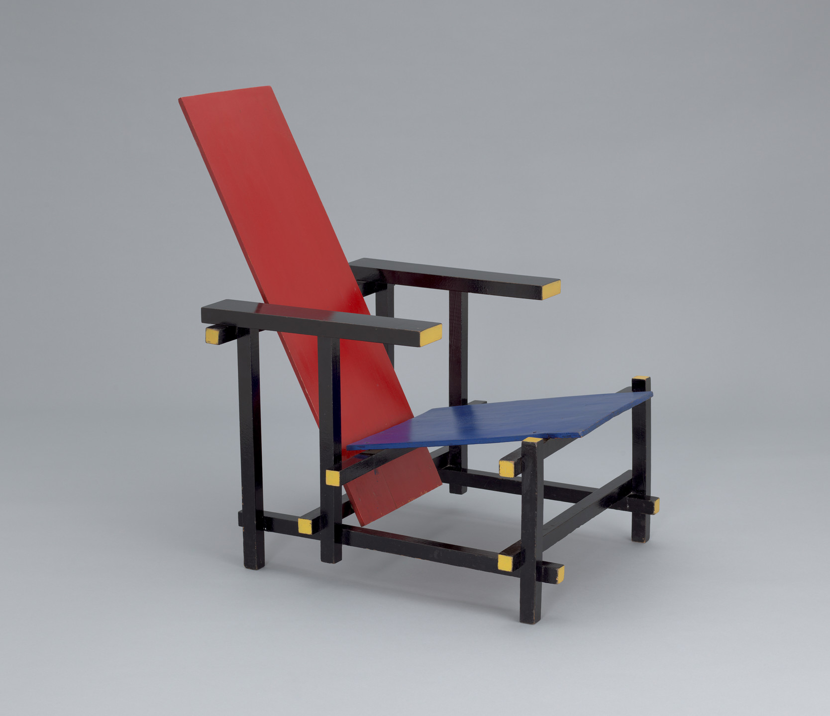 A chair with a red backrest, a blue seat, and a black frame with yellow accents on a grey background. The chair has wooden arms and legs structured with straight lines and right angles. This is an AI generated description. To give feedback, email ai@moma.org.