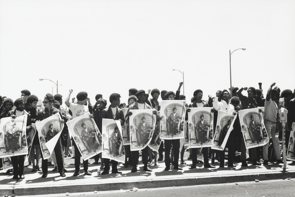 Black Panthers march in formation at the Alameda County Courthouse. They wear black leather jackets, berets. Many have raised fists. They carry identical posters showing Huey P. Newton, seated in a wicker chair, holding a rifle and a spear. The posters read FREE HUEY. The image is bright, sun-bleached, almost overexposed. The street stretches back, lined with lamp posts.