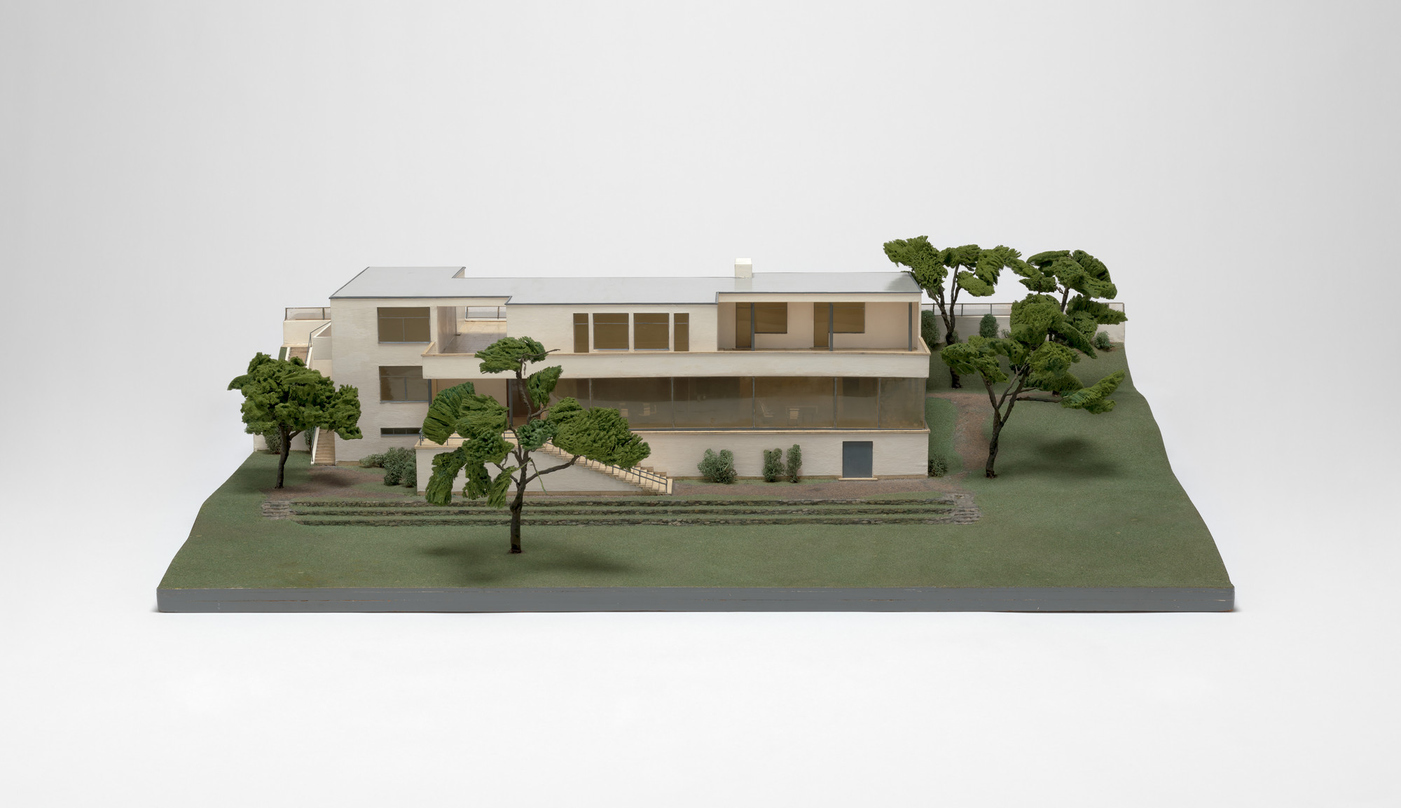 A scale model of a two-story rectangular white building with large windows, surrounded by green trees and grass. The building has stairs on one side and a flat roof. This is an AI generated description. To give feedback, email ai@moma.org.