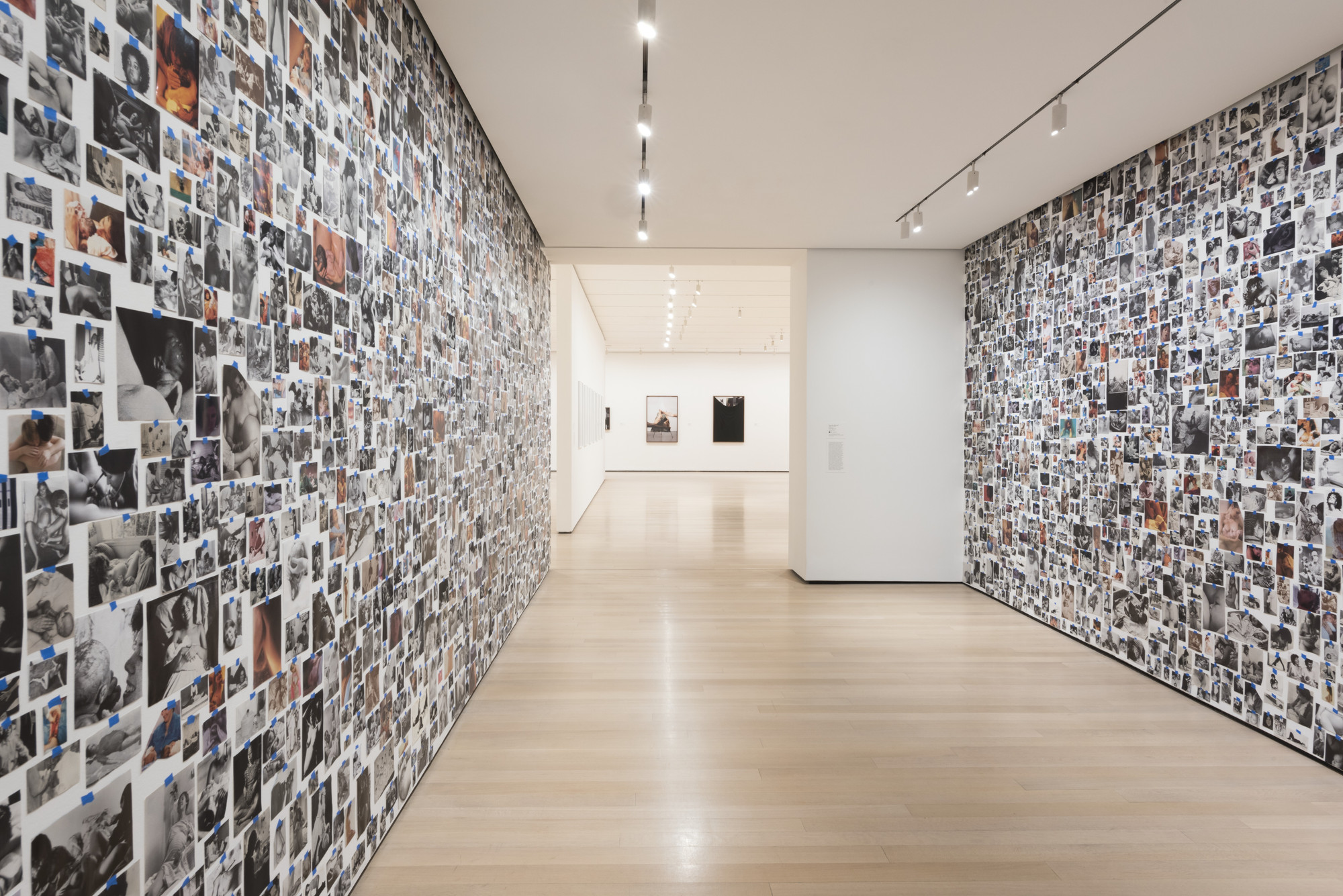 A hallway with light wood floors is lined with hundreds of black-and-white and colored photos covering both walls. The ceiling has rows of spotlights, and the hallway opens into a white-walled room with framed artworks. This is an AI generated description. To give feedback, email ai@moma.org.