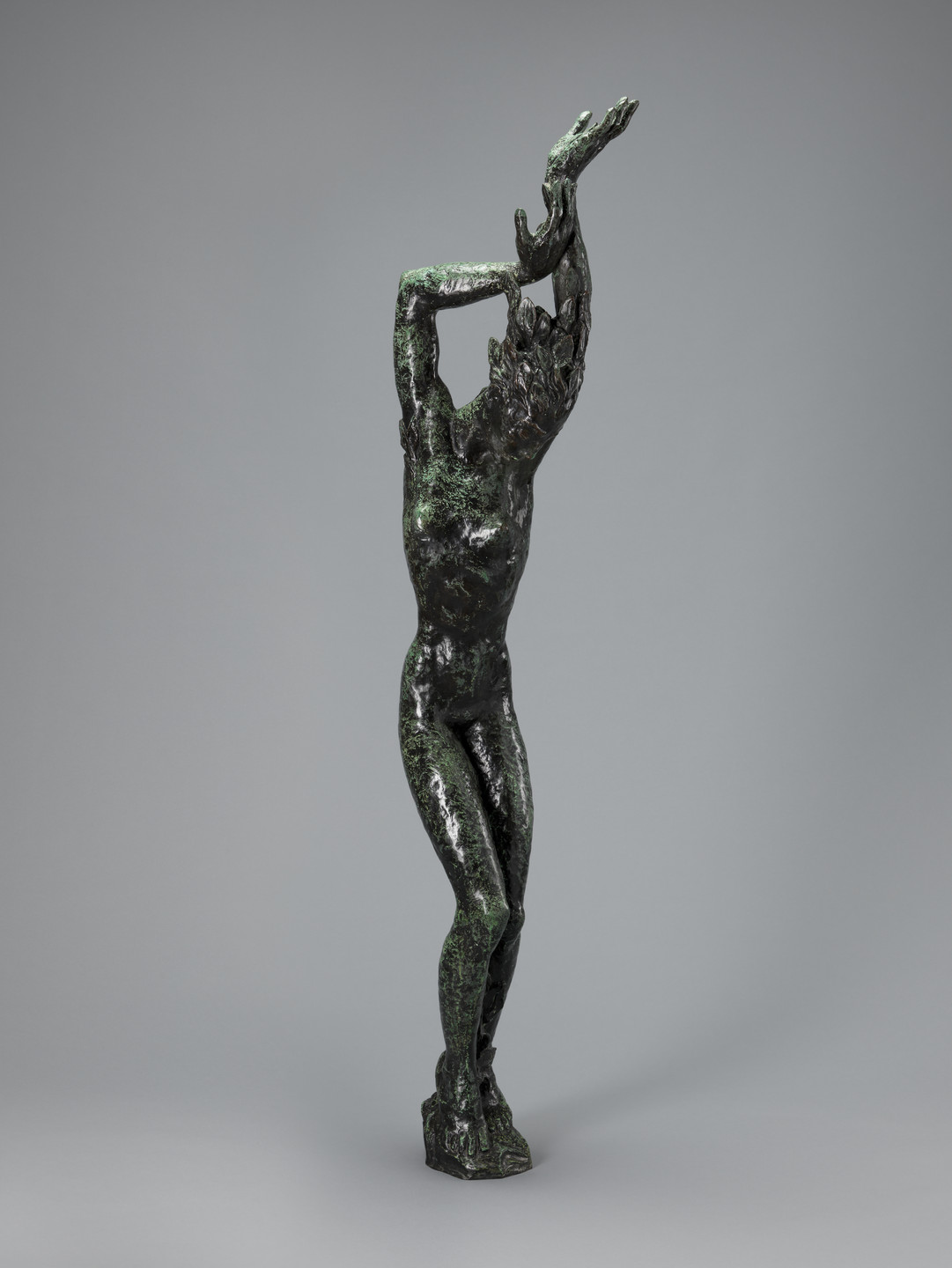 A dark green sculpture of a standing human figure with arms raised, one hand touching the opposite arm above. The figure&#39;s legs are crossed at the ankles, and the background is plain gray. This is an AI generated description. To give feedback, email ai@moma.org.