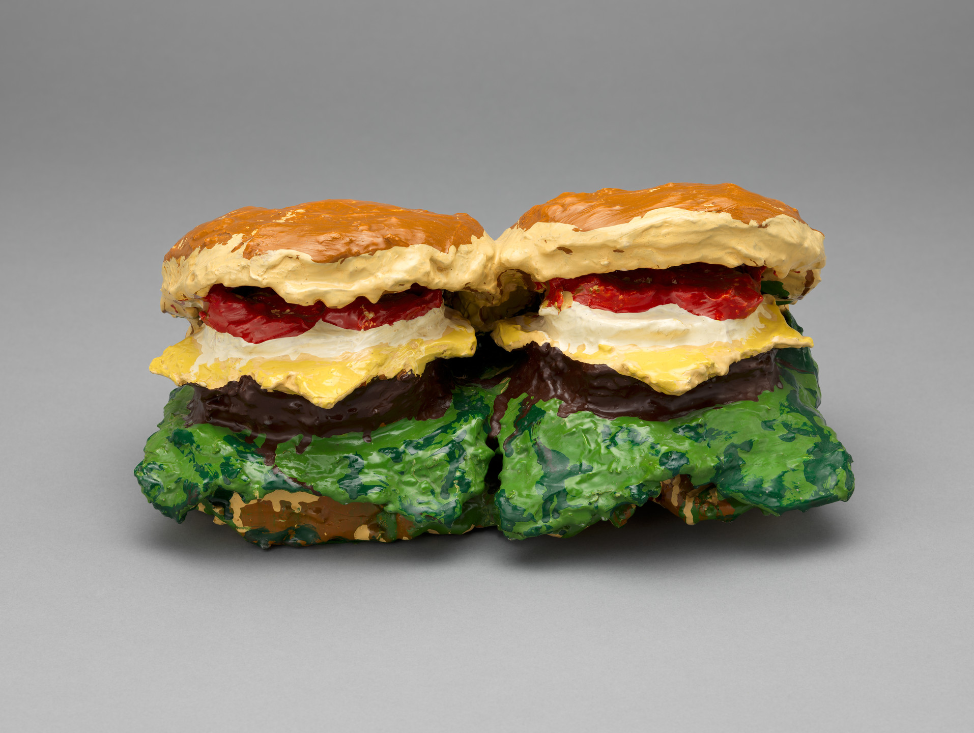 The image shows a painted sculpture of two burgers side by side, with many layers. The colors used are predominantly brown for the buns, green for the lettuce, red for tomatoes, yellow for cheese, and white for an additional layer. This is an AI generated description. To give feedback, email ai@moma.org.