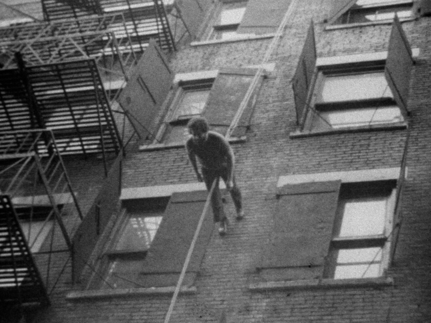 A person is suspended by a rope, mid-air, in front of a brick building with several open windows. The building has a fire escape on the left side with metal stairs and railings. This is an AI generated description. To give feedback, email ai@moma.org.
