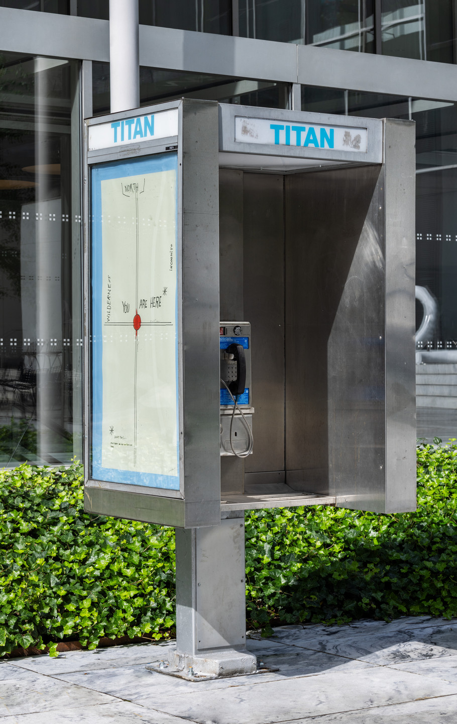 A metallic phone booth stands outdoors with greenery at its base. Inside, a blue payphone is visible, and one side features a map labeled &#34;You are here&#34; with a red dot. This is an AI generated description. To give feedback, email ai@moma.org.
