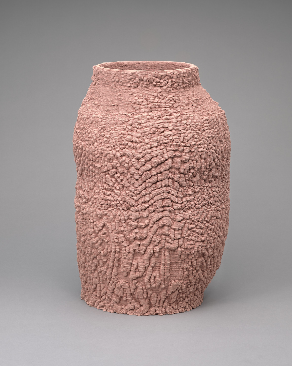 A tall, textured vase in a dusty pink color stands against a light gray background. The vase&#39;s surface has an irregular, bumpy pattern, creating a tactile, almost rugged appearance. This is an AI generated description. To give feedback, email ai@moma.org.