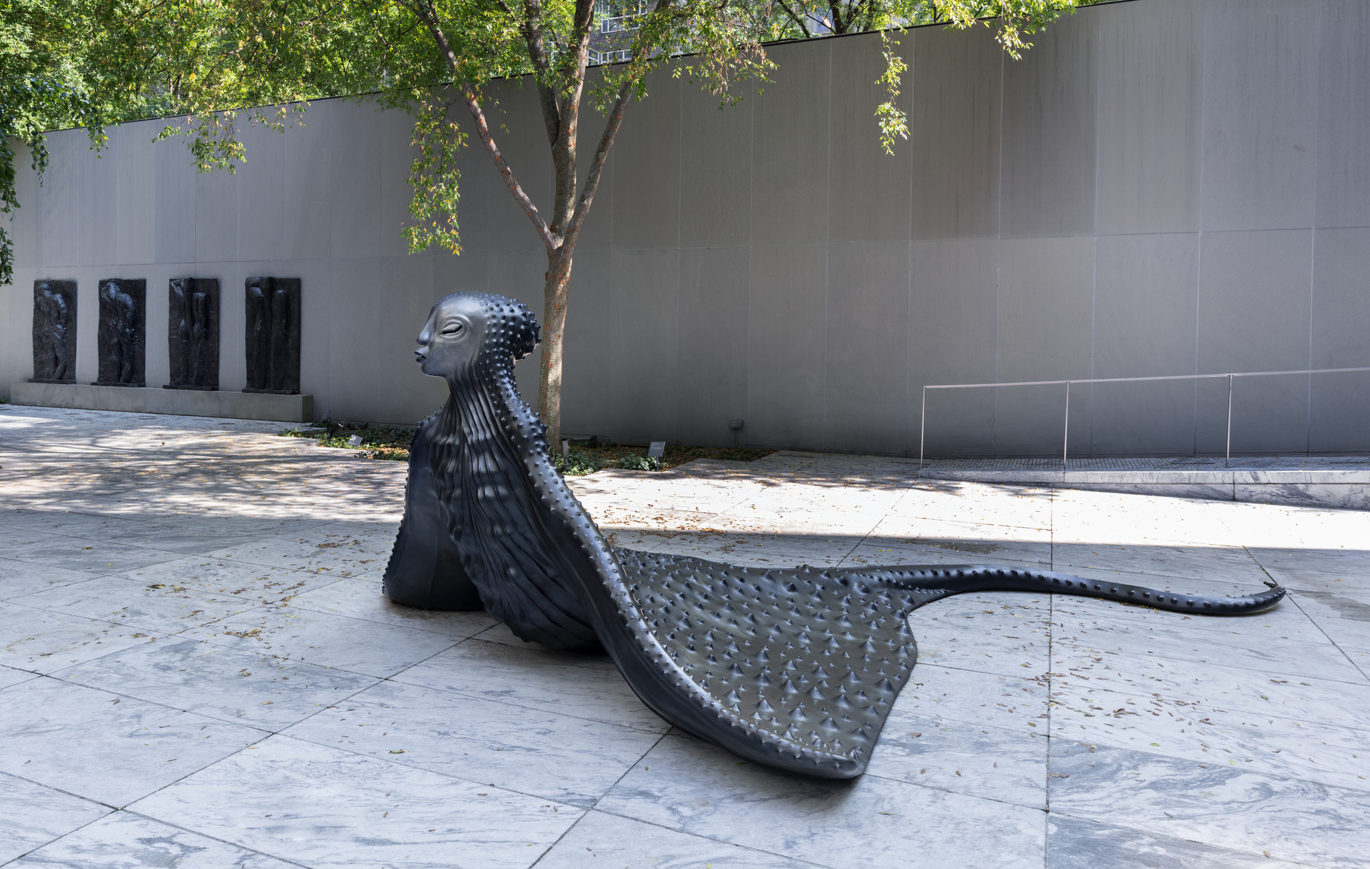 A dark grey sculpture resembling a human-headed creature with a long, textured tail is placed on a light grey marble ground. Behind it, there is a grey wall, trees, and four vertical, dark stone panels. This is an AI generated description. To give feedback, email ai@moma.org.