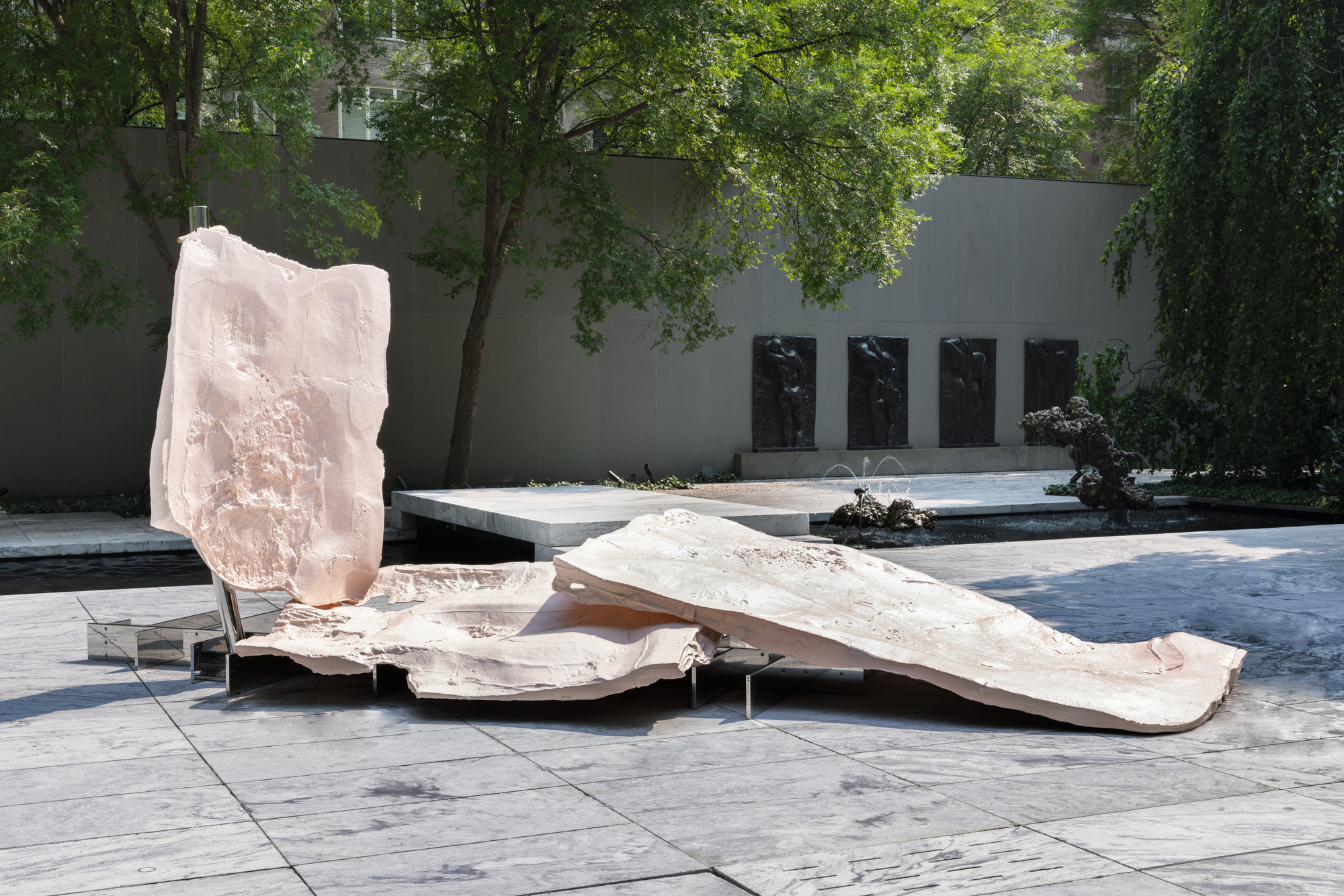 A large pink sculptural piece with rough textures is set on a gray stone floor in an outdoor space, surrounded by green trees. In the background is a gray wall with dark reliefs and a small fountain. This is an AI generated description. To give feedback, email ai@moma.org.
