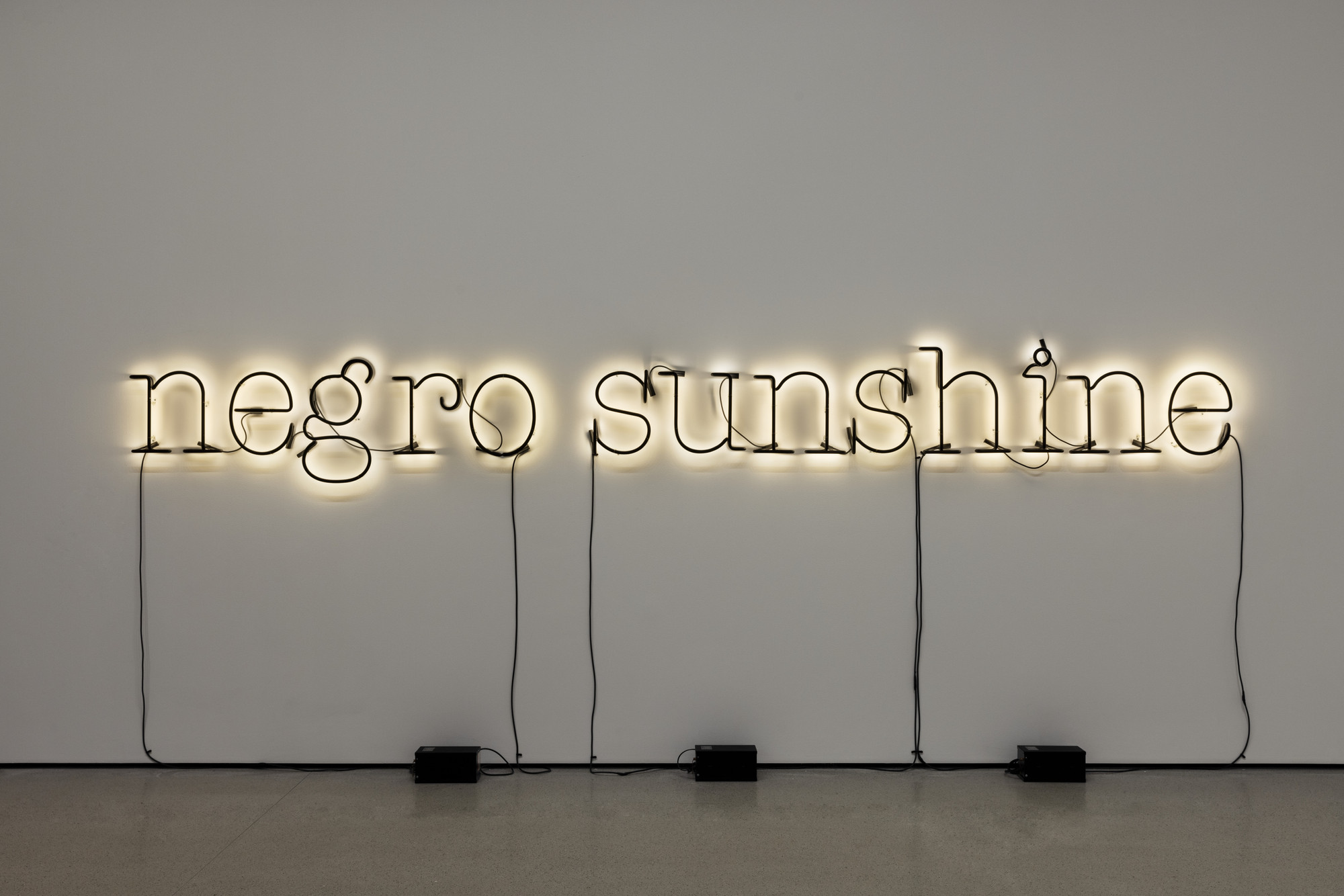 A neon light sculpture on a white wall spells out the words "negro sunshine" in black cursive with a yellow glow. Black cords run from the letters to power sources on the floor. This is an AI generated description. To give feedback, email ai@moma.org.