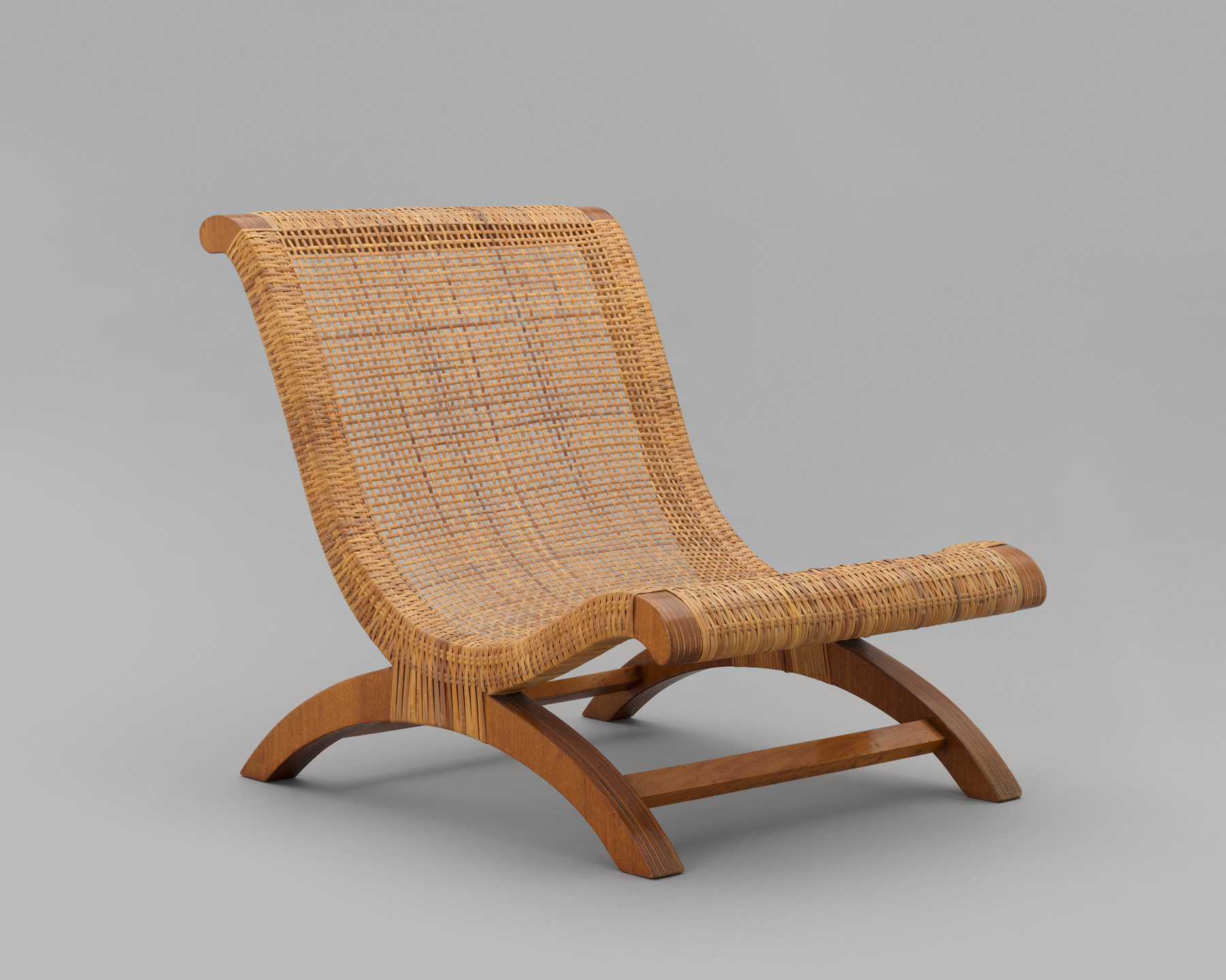 A wooden chair features a low, curved seat and backrest made of woven light brown material. The chair's legs and frame are smooth, dark brown wood, forming an arc shape. This is an AI generated description. To give feedback, email ai@moma.org.