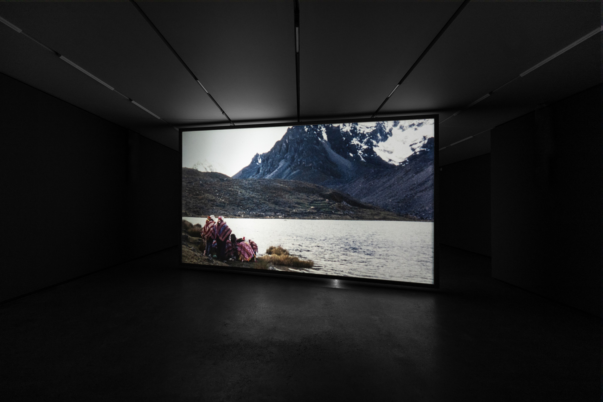 A large screen in a dark room displays a photograph of people near a lake with snow-capped mountains in the background. The colors predominantly include dark shades, white, and muted browns with some red. This is an AI generated description. To give feedback, email ai@moma.org.