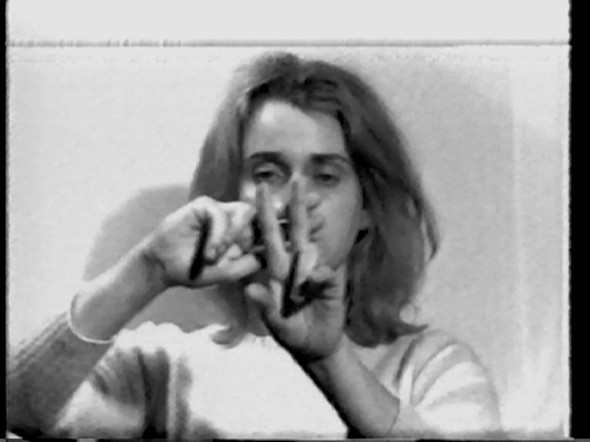 A grayscale image of a person, with shoulder-length hair, gesturing with their hands in front of their face. They are wearing a light-colored long-sleeve shirt and are positioned against a plain background. This is an AI generated description. To give feedback, email ai@moma.org.