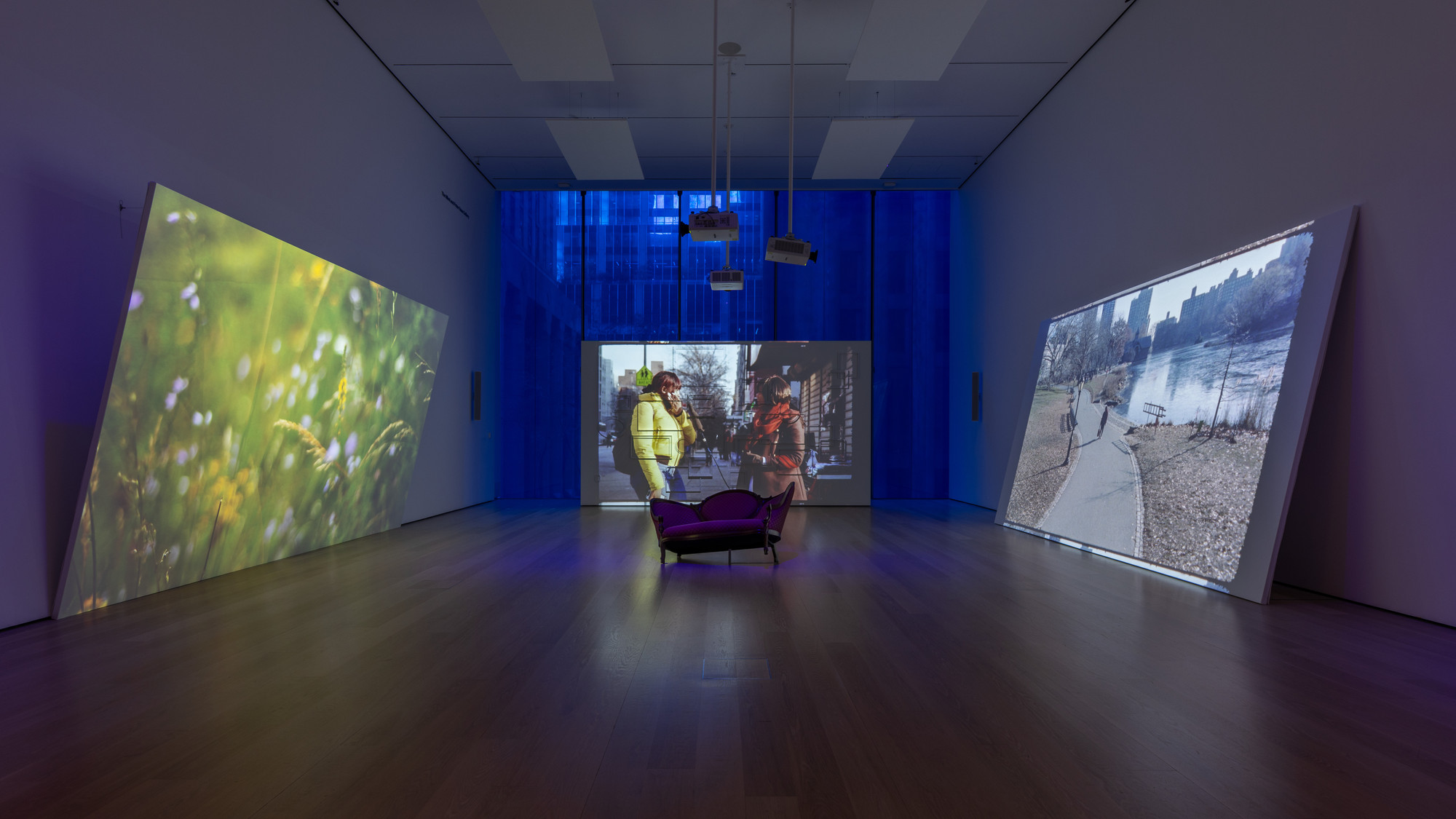 The room has three large screens displaying different scenes: a green field with white flowers, two people conversing, and a riverside walkway. A purple couch is centered in front of the middle screen, and blue lighting fills the room. This is an AI generated description. To give feedback, email ai@moma.org.