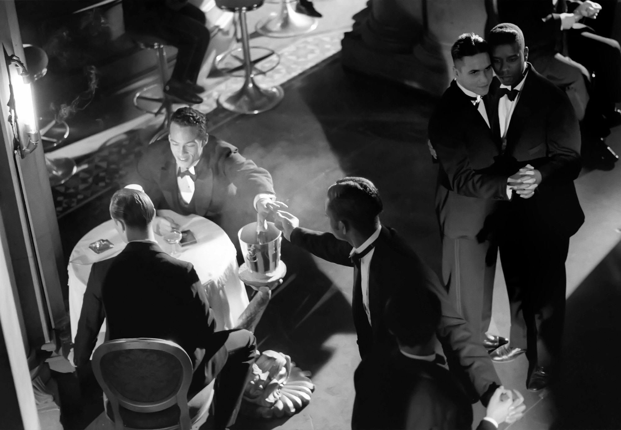 In a dimly lit room, people dressed in dark suits and bow ties are engaged in various activities; two are seated at a table, while others are standing and dancing. The image is primarily in shades of black and white, creating a vintage feel. This is an AI generated description. To give feedback, email ai@moma.org.