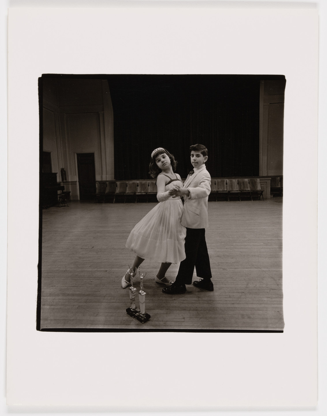 Two children in formal attire dance in the center of an empty hall with wooden floors; two trophies are placed at their feet. The background features rows of chairs and a dark curtain. This is an AI generated description. To give feedback, email ai@moma.org.