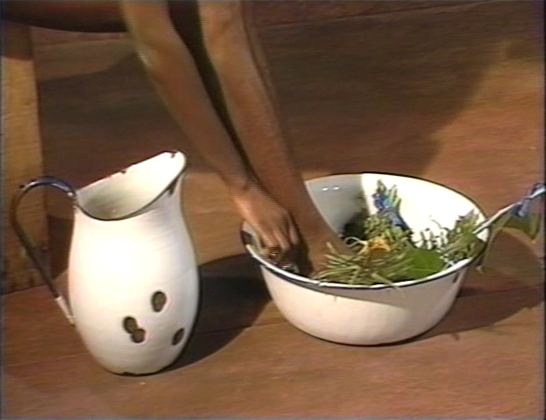 A white jug with black spots is placed next to a white bowl containing green leafy plants. A person&#39;s arms are reaching into the bowl, which is set on a brown wooden surface. This is an AI generated description. To give feedback, email ai@moma.org.