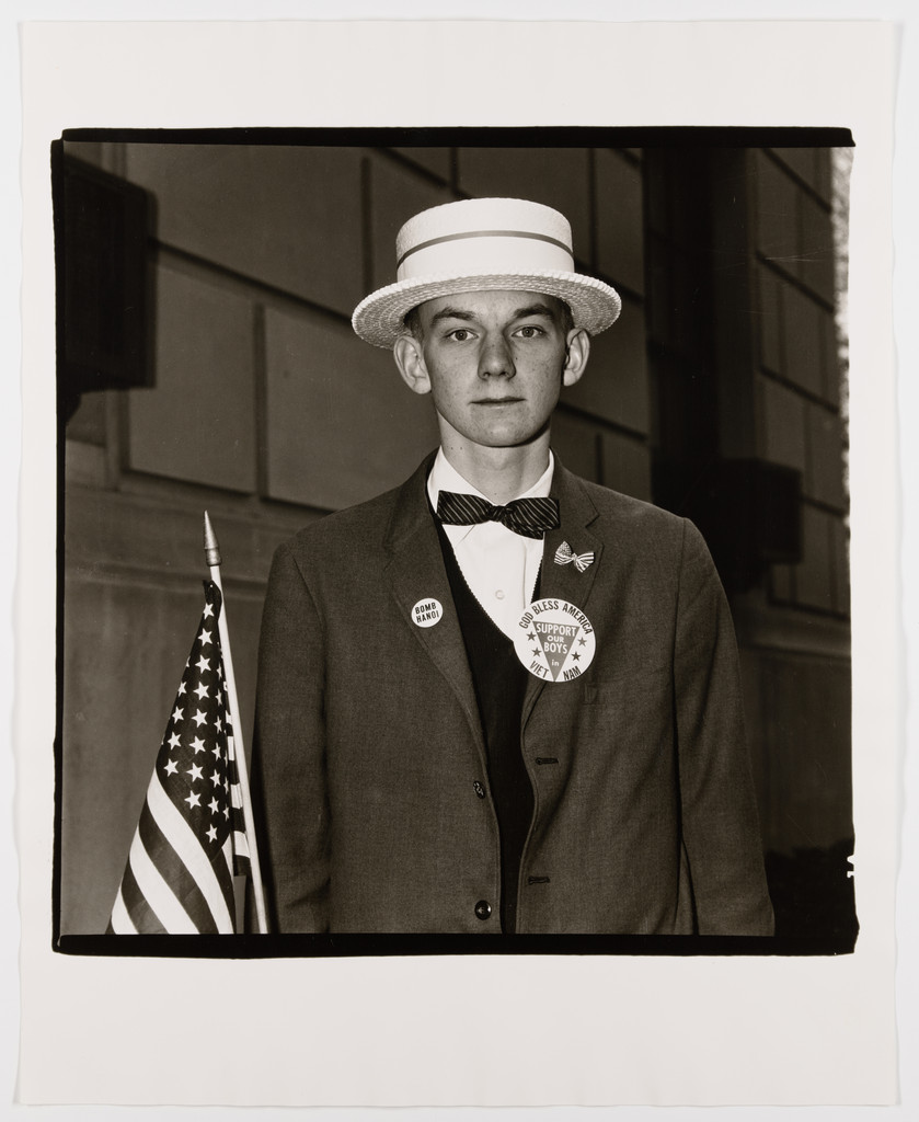 A young man in a straw boater hat, bow tie, and dark suit stares directly at the camera. He holds a small American flag and wears buttons reading 'Bomb Hanoi' and 'God Bless America / Support Our Boys in Viet Nam.' Behind him, a stone building. Diane Arbus's characteristic square format with irregular black borders frames the image.