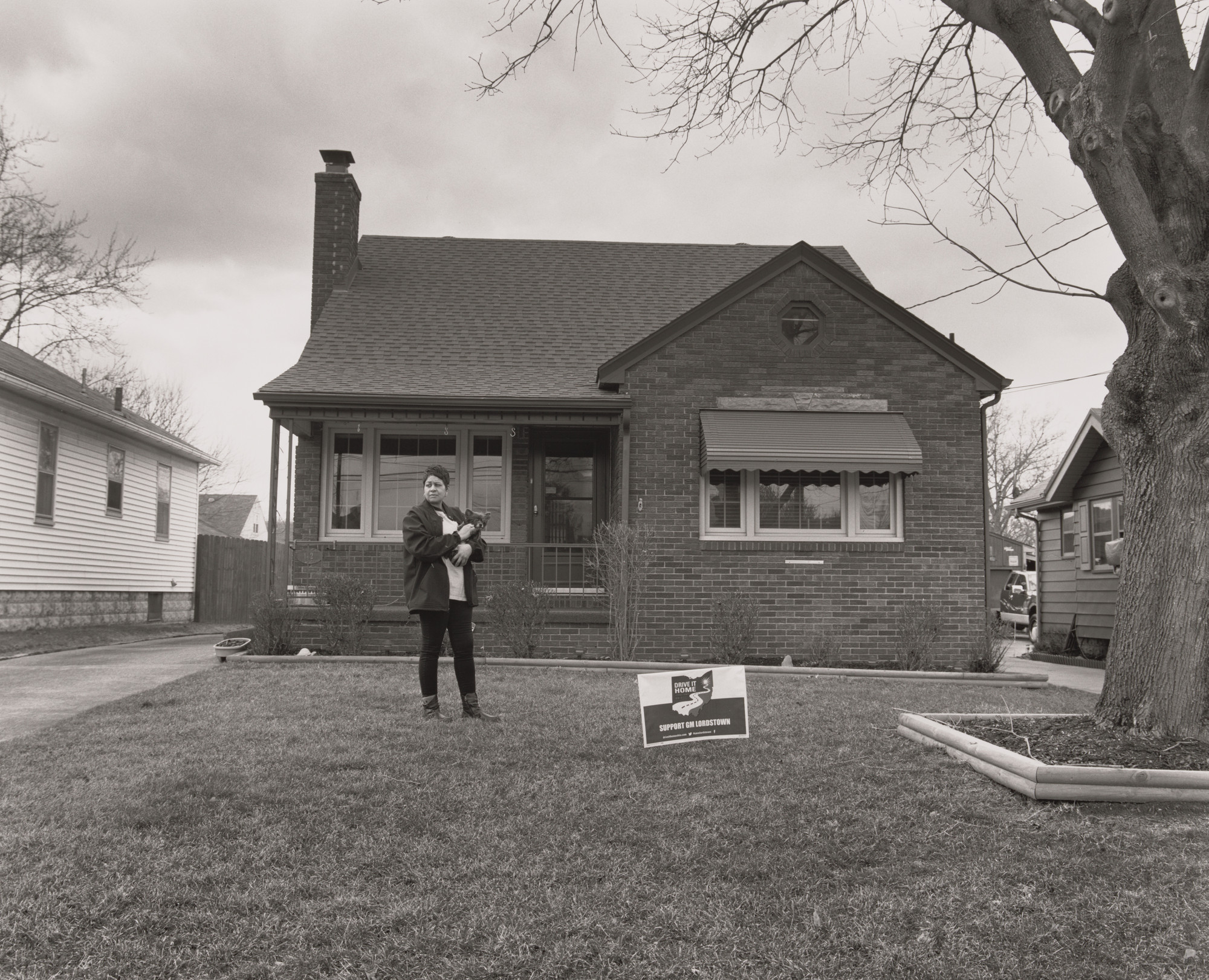 LaToya Ruby Frazier. Kesha Scales, UAW Local 1714, standing on her front lawn with a Drive-It-Home Campaign sign and holding her dog Gigi, (22 years in at GM Lordstown Complex, pressroom), Youngstown, OH.