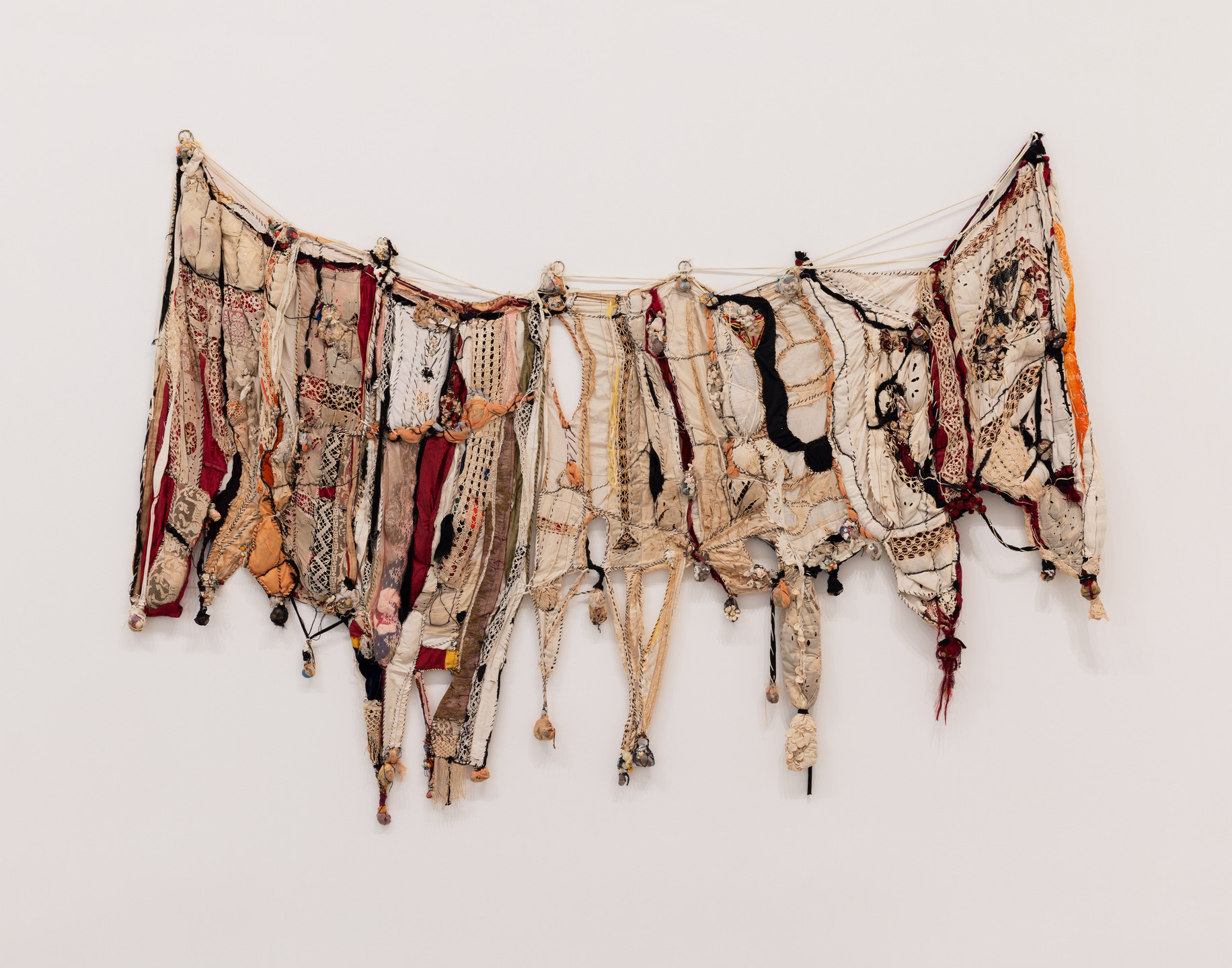 A textile hanging on the wall features an array of neutral and earthy tones, with predominant colors being beige, brown, red, and hints of black. It consists of various fabric strips and patterns woven together with some hanging threads. This is an AI generated description. To give feedback, email ai@moma.org.