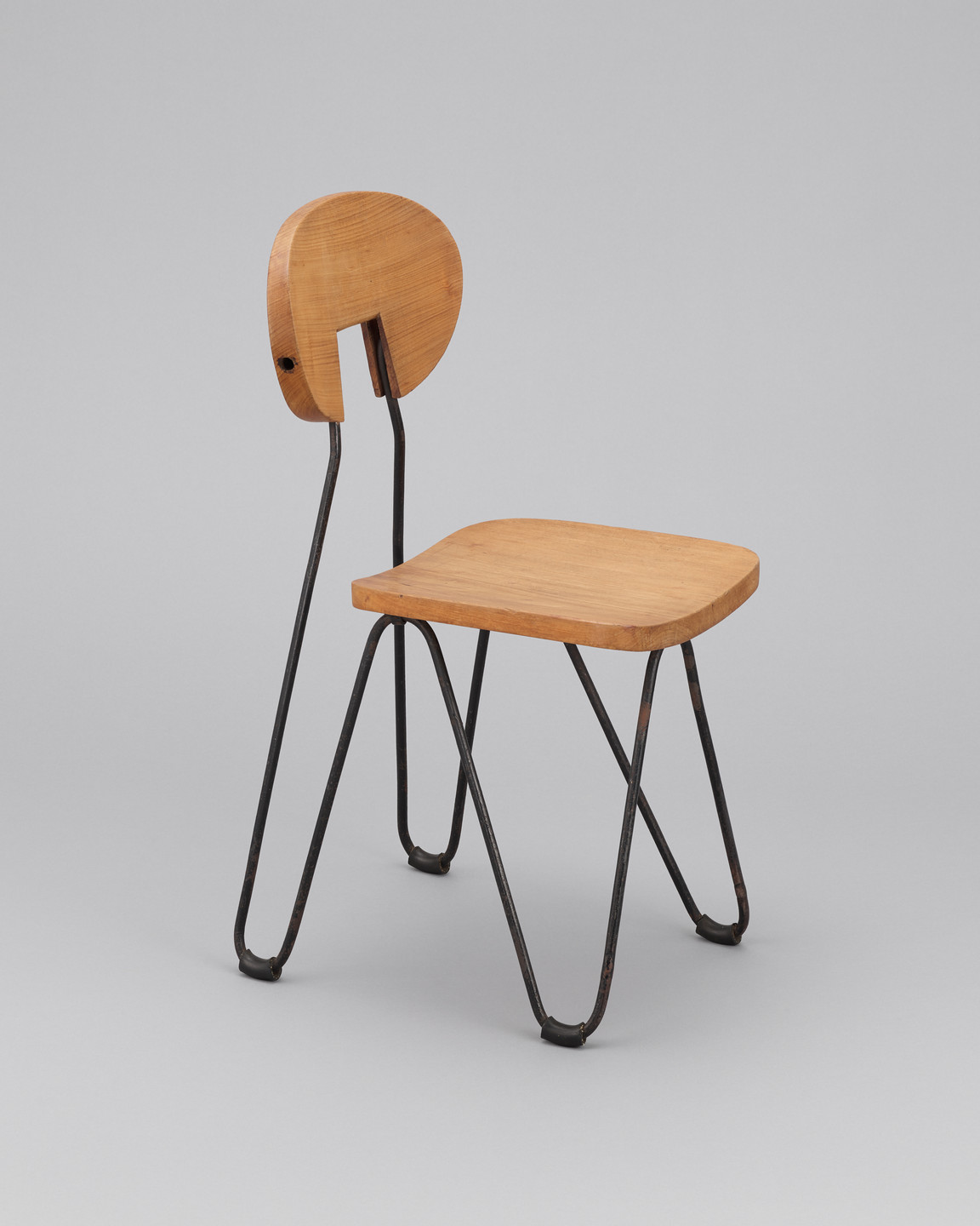 A wooden chair with a flat seat, rounded back with a gap, and thin black metal legs. The background is plain gray. This is an AI generated description. To give feedback, email ai@moma.org.