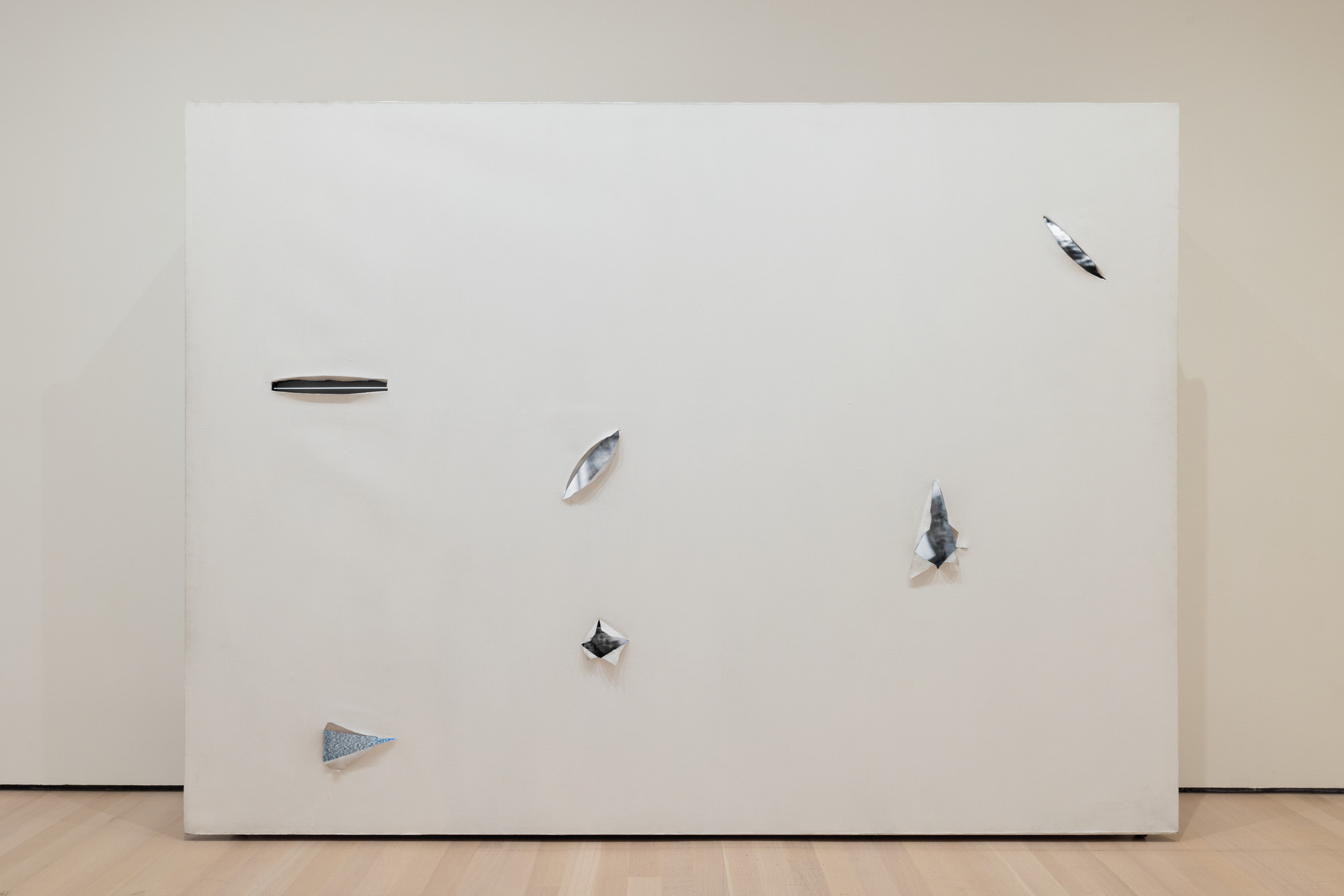 A large white canvas features six irregular, shiny silver shapes distributed unevenly across its surface. The canvas is displayed in a room with light wooden flooring and beige walls. This is an AI generated description. To give feedback, email ai@moma.org.
