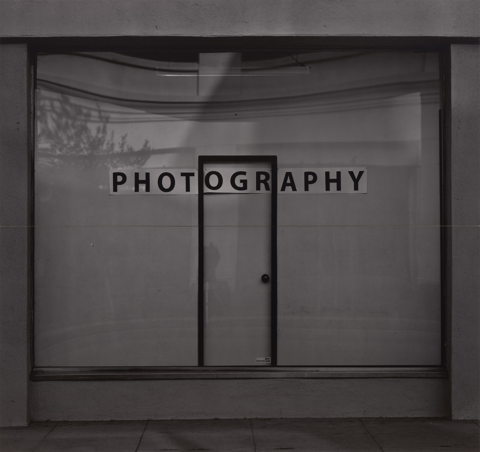 A grayscale image features a storefront with a glass door and large window, with the word &#34;PHOTOGRAPHY&#34; in bold, black letters above the door. Shadows of tree branches are visible on the glass. This is an AI generated description. To give feedback, email ai@moma.org.