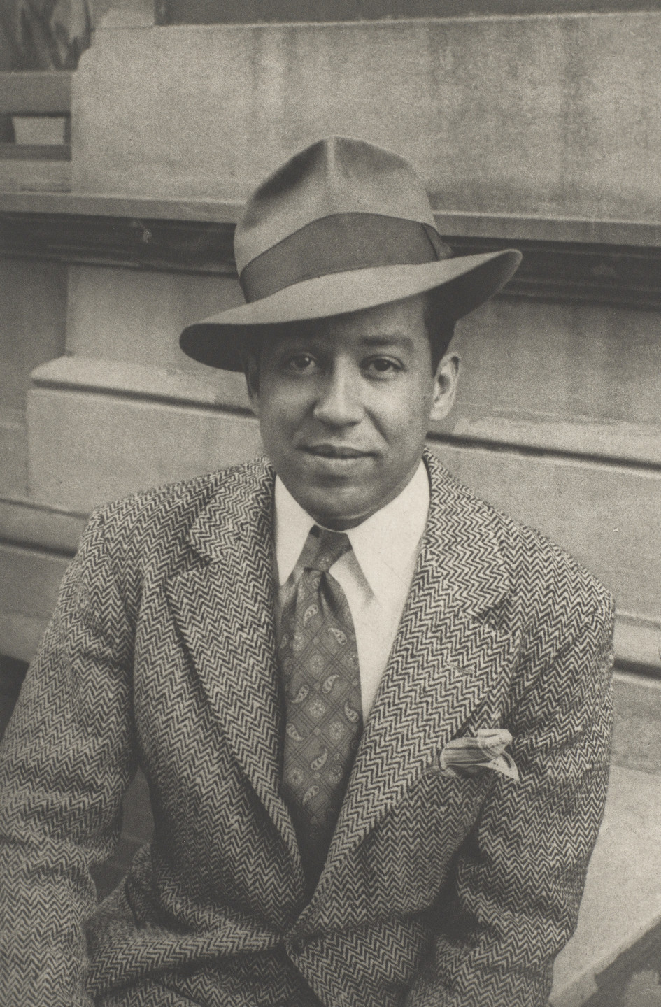 A person is sitting in front of a stone building, wearing a patterned suit, a paisley tie, and a tilted fedora. The image is in black and white. This is an AI generated description. To give feedback, email ai@moma.org.