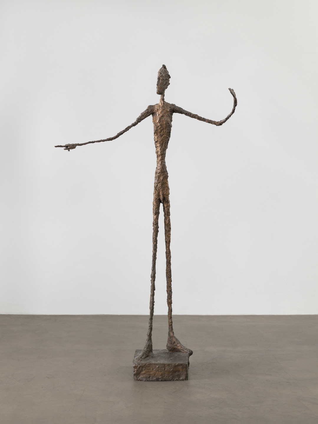 A tall, bronze-colored sculpture depicts an elongated human figure standing on a rectangular base. The figure has thin limbs, an elongated torso, and outstretched arms. This is an AI generated description. To give feedback, email ai@moma.org.