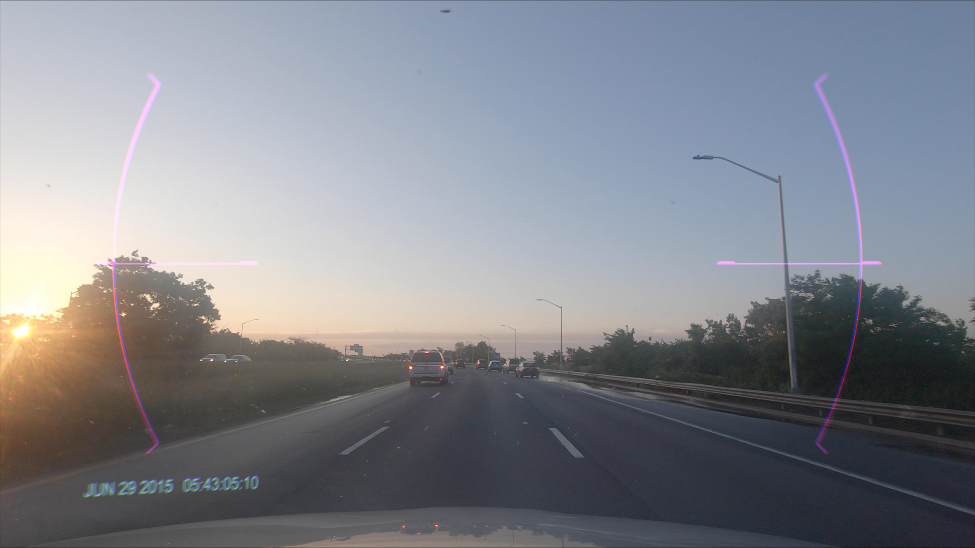 A road scene during sunrise with the sun low on the horizon, trees on the left, and cars on the road heading into the distance. The sky is mostly clear with soft shades of blue and orange hues from the sunrise. This is an AI generated description. To give feedback, email ai@moma.org.