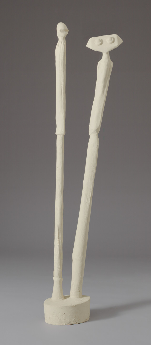 A cream-colored sculpture depicts two elongated, textured figures standing side by side on a circular base against a gray background. The taller figure on the left has a round head, while the shorter figure on the right has an angular head with two raised circular shapes resembling eyes. This is an AI generated description. To give feedback, email ai@moma.org.