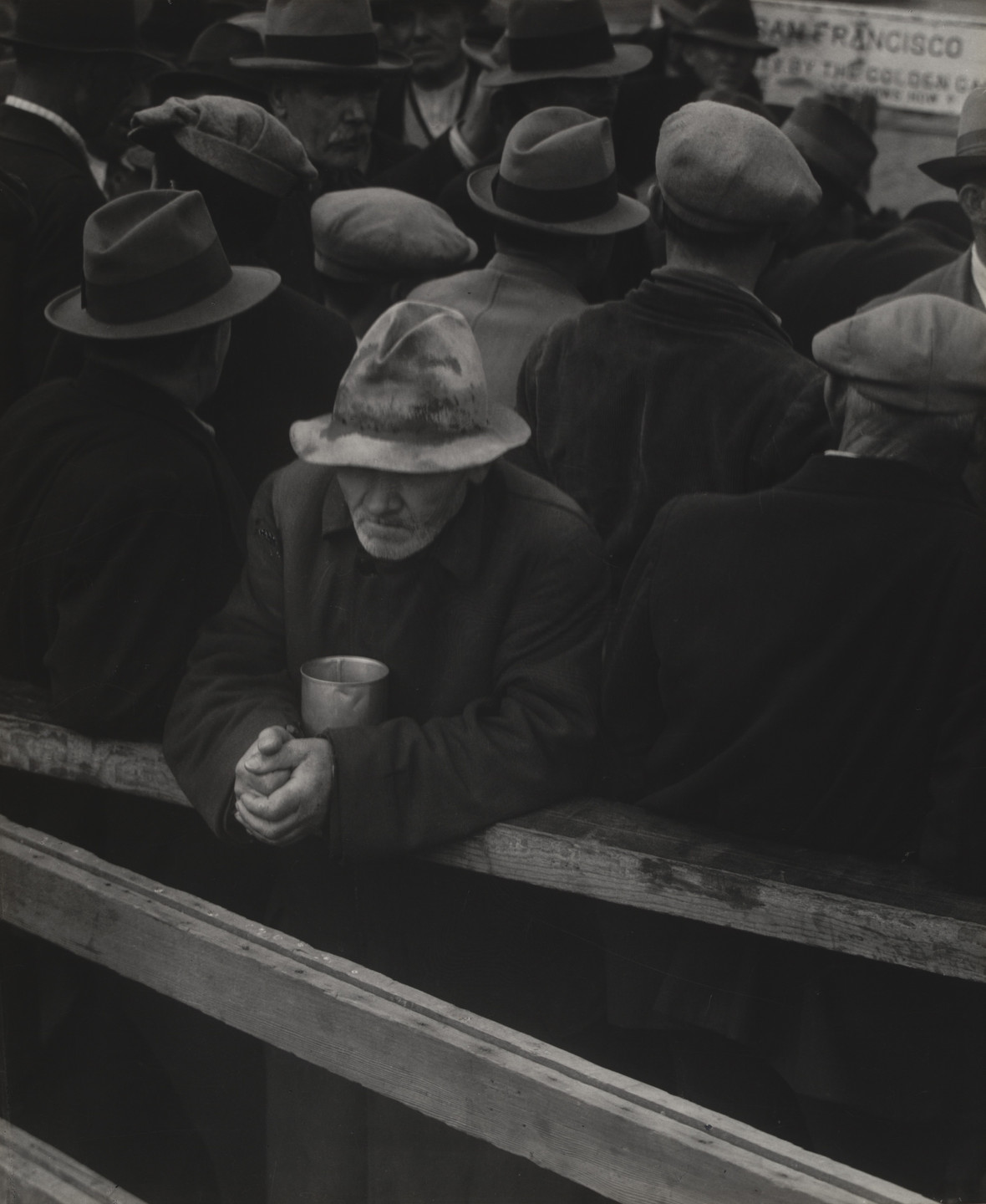 A person in a worn hat and dark coat holds a metal cup and leans on a wooden barrier, surrounded by others wearing hats and coats. The image is in grayscale, creating a somber atmosphere. This is an AI generated description. To give feedback, email ai@moma.org.