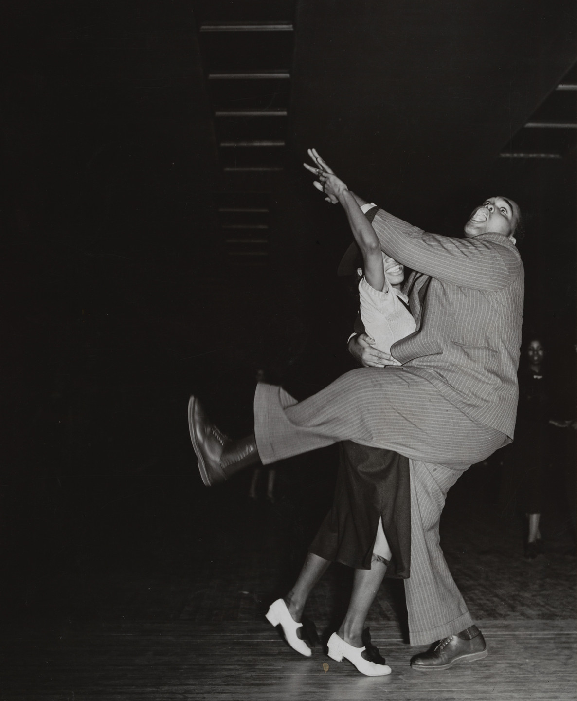 Two people are dancing energetically indoors; one person is lifted in the air, with legs bent and arms reaching out. The image is in black and white, with a dark background. This is an AI generated description. To give feedback, email ai@moma.org.