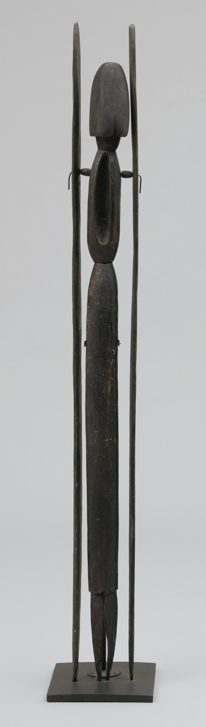 A tall, slender wooden sculpture with a dark brown, weathered finish, featuring an elongated oval head, elongated body, and rectangular base. The figure has thin protruding side elements. This is an AI generated description. To give feedback, email ai@moma.org.