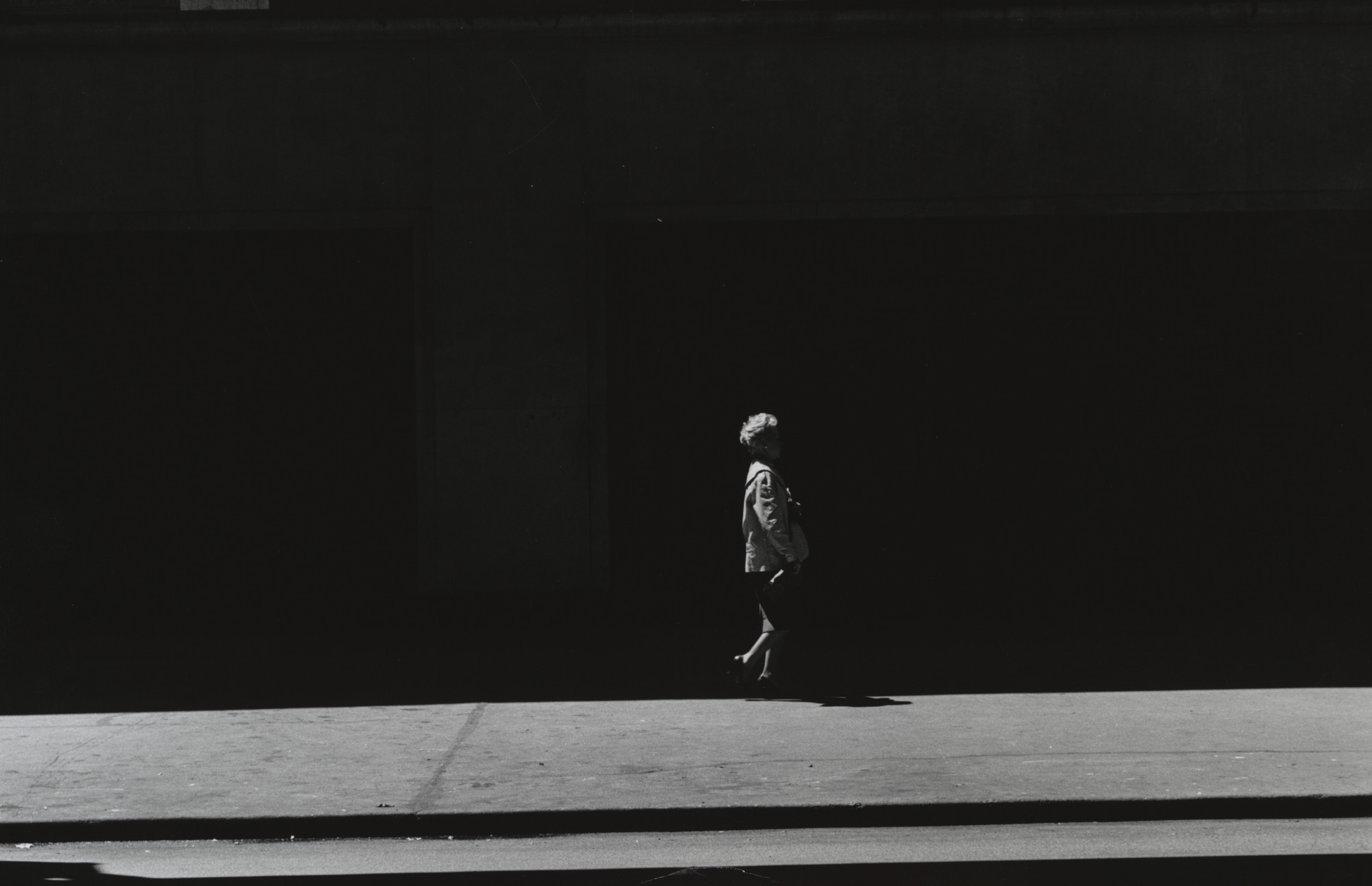 A person wearing a light-colored jacket is walking on a sunlit sidewalk in front of a dark building. The predominant colors are black and white, with strong shadows creating contrast. This is an AI generated description. To give feedback, email ai@moma.org.