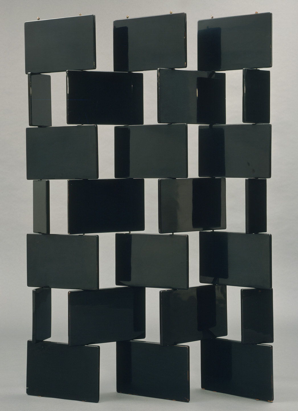 The image shows a vertical arrangement of dark rectangular panels with gaps between them, creating a staggered pattern. The background is light grey, enhancing the contrast of the dark panels. This is an AI generated description. To give feedback, email ai@moma.org.