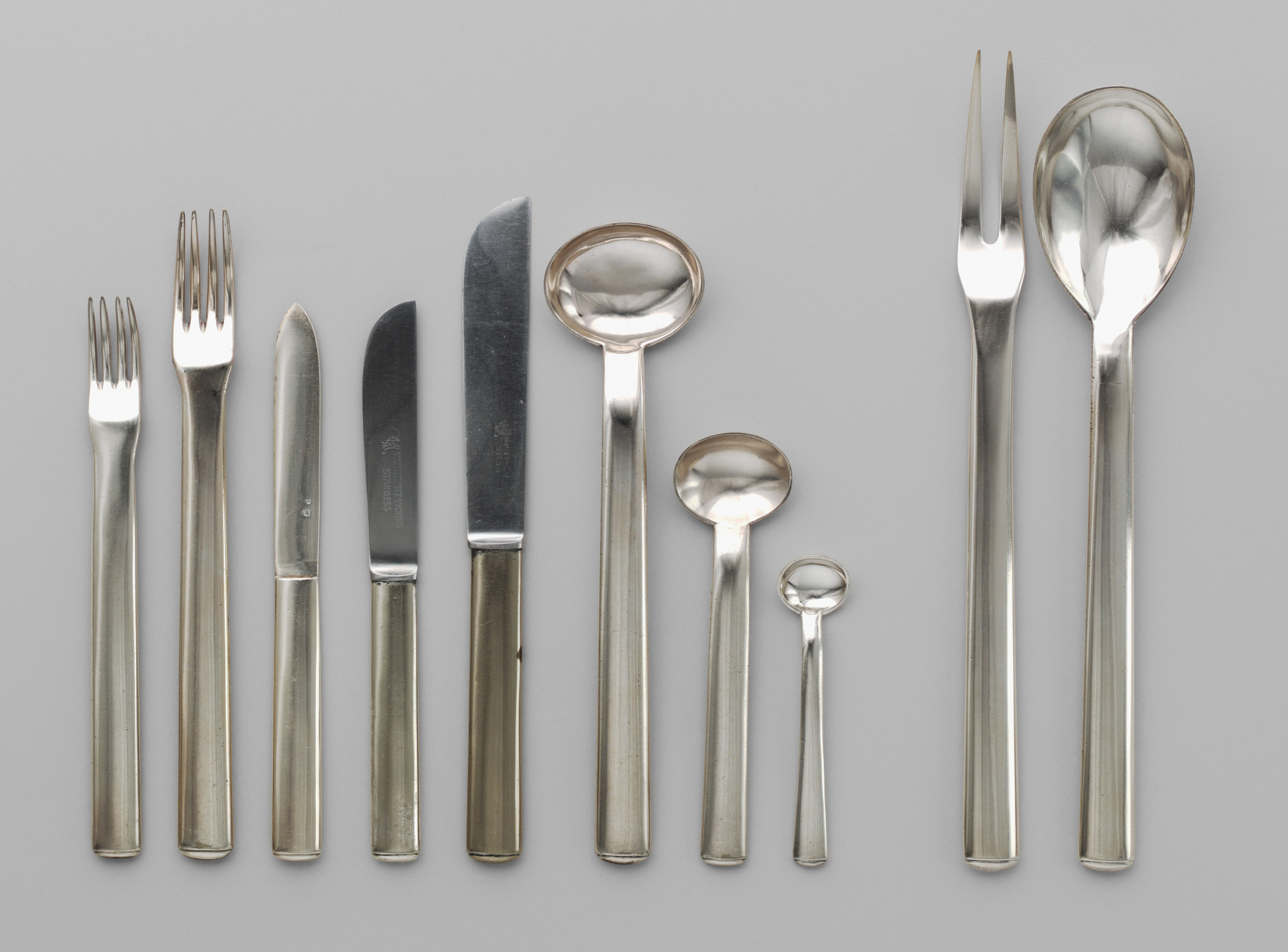 A set of nine silver utensils, including three forks, two knives, three spoons, and one serving fork, are arranged in ascending size on a light gray background. The utensils have a reflective, metallic finish. This is an AI generated description. To give feedback, email ai@moma.org.