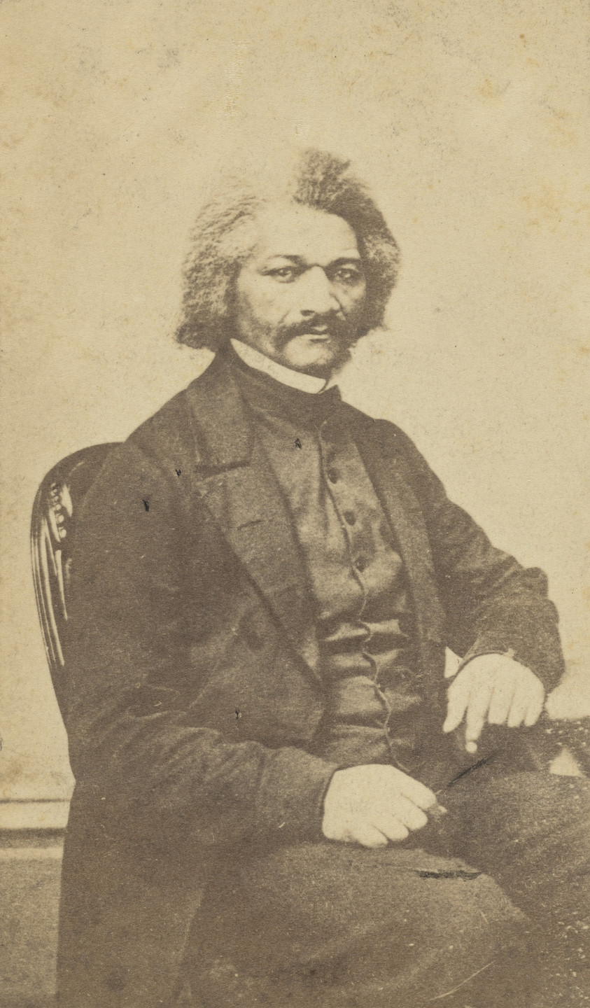 A person with short, curly hair and a mustache, dressed in a dark suit, sits on a chair, facing the camera. The photograph has a sepia tone, giving it a warm, brownish color. This is an AI generated description. To give feedback, email ai@moma.org.
