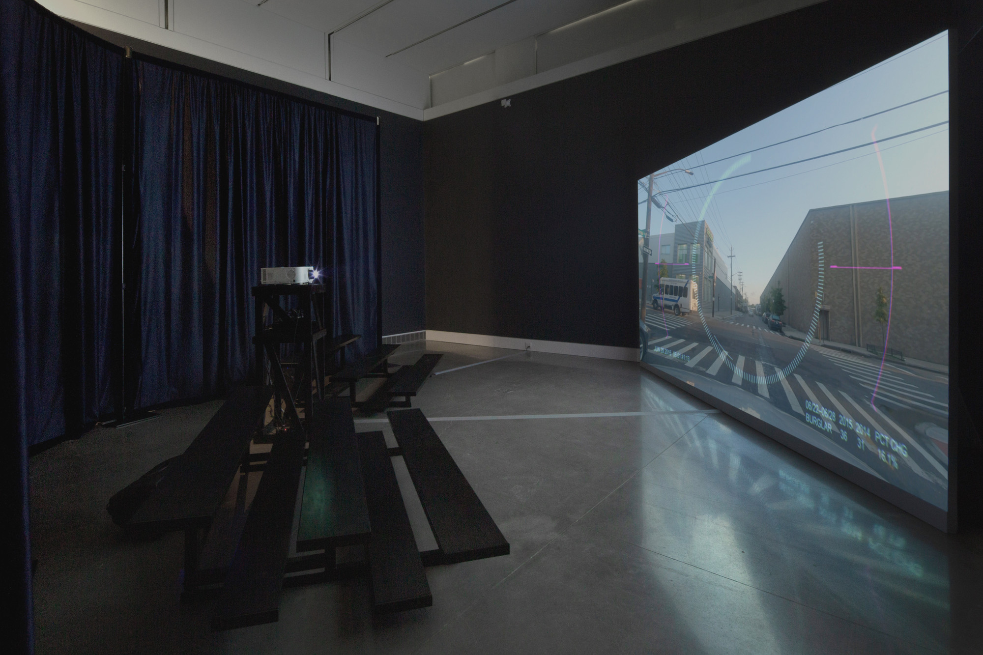 A dark room with black benches faces a large screen displaying a city street scene with buildings, power lines, and a bus. A projector on a stand is positioned near the benches. This is an AI generated description. To give feedback, email ai@moma.org.