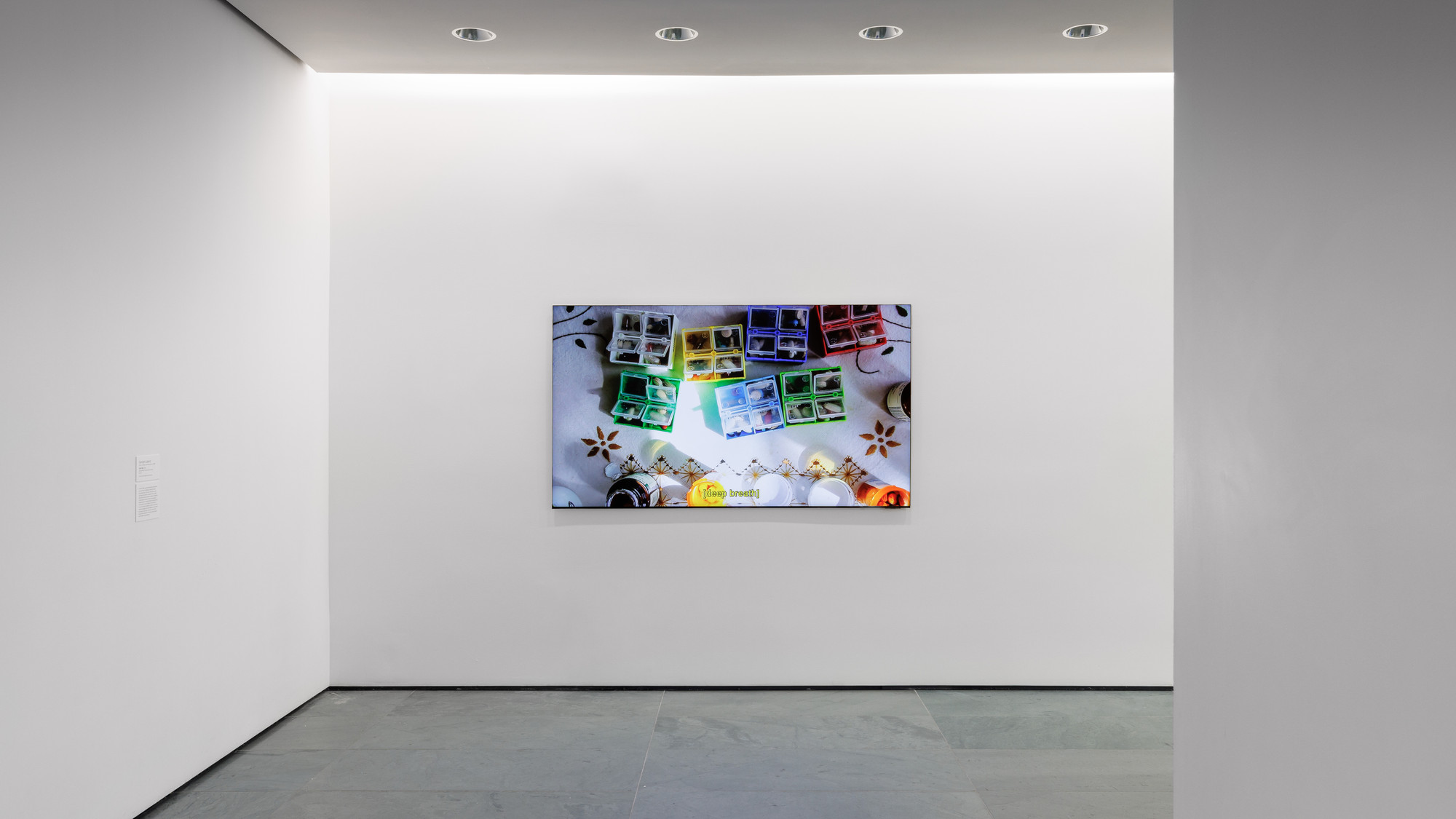 The image shows a brightly lit art gallery with a large screen on the wall displaying colorful compartments holding various items. The predominant colors are white, green, blue, and yellow. This is an AI generated description. To give feedback, email ai@moma.org.