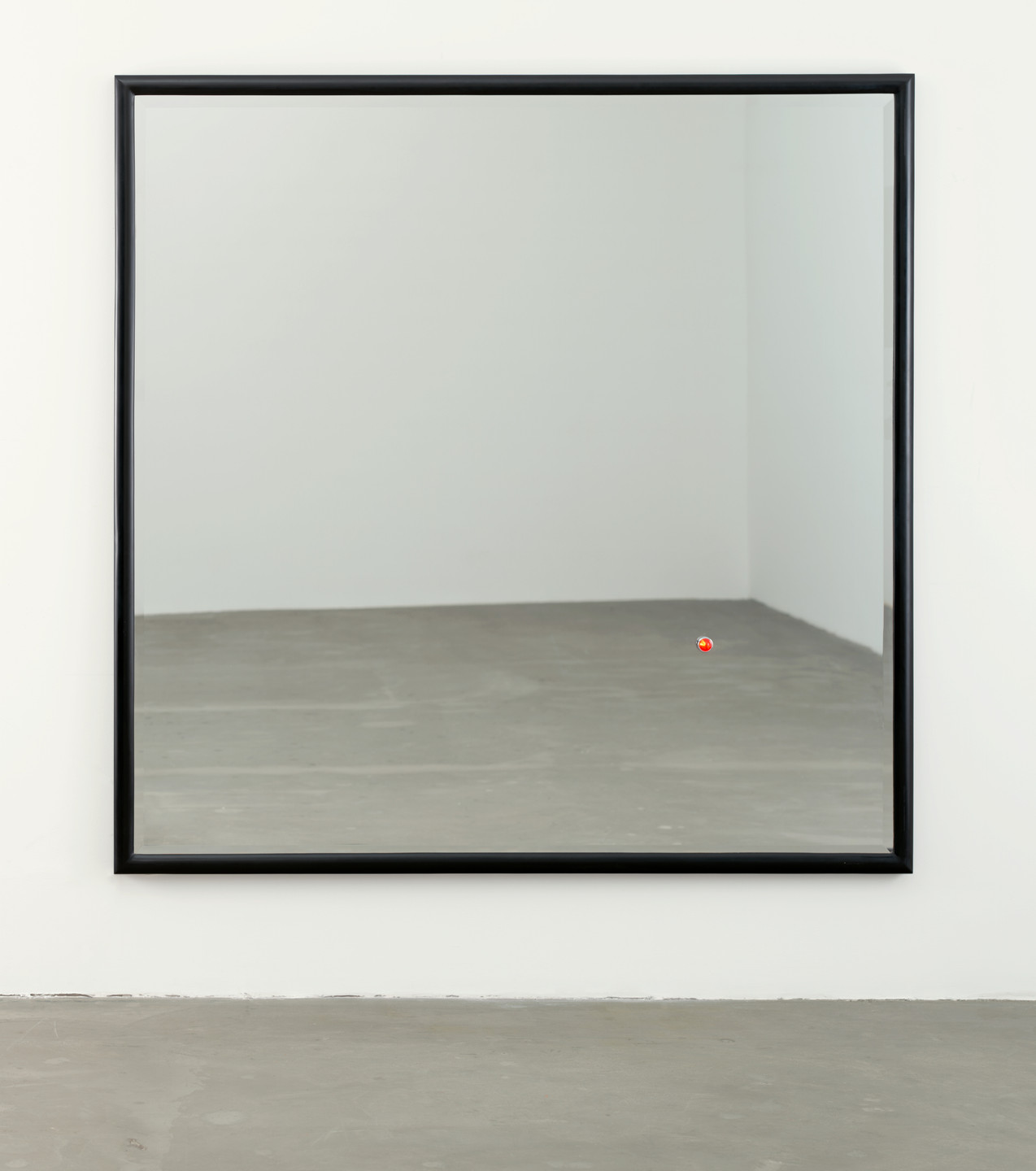 A large mirror with a black frame reflects a room with gray floors and white walls. A small red sphere is visible on the bottom right corner of the mirror. This is an AI generated description. To give feedback, email ai@moma.org.