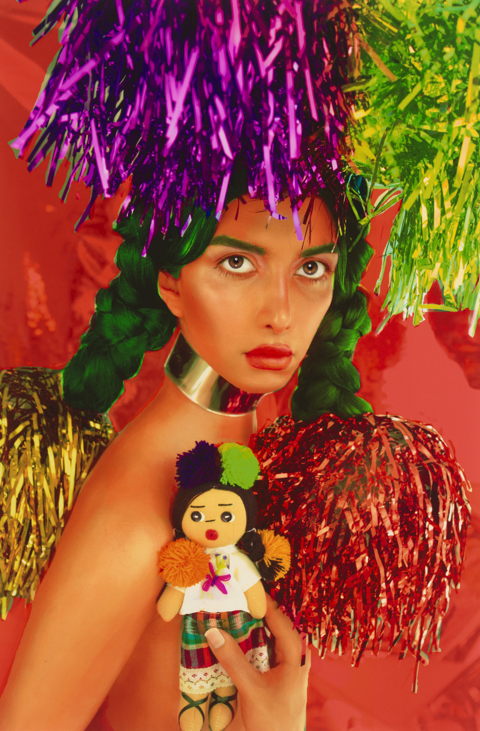 A person with braided green hair holds a small doll, surrounded by colorful tinsel in purple, green, and red. The background is vibrant red, and the person wears a metallic choker. This is an AI generated description. To give feedback, email ai@moma.org.