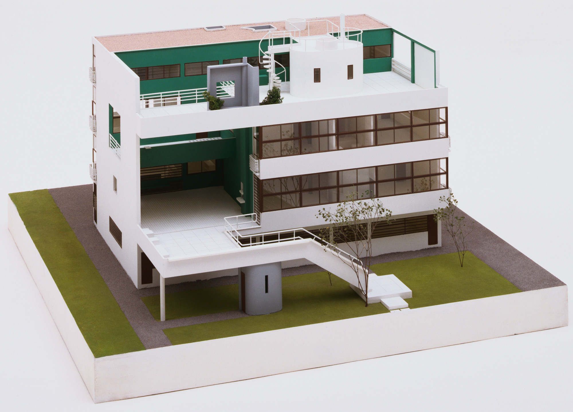 A model of a multi-story building with white walls, green sections, and large windows. The building has a rooftop terrace, exterior stairs, and small trees in the surrounding grassy area. This is an AI generated description. To give feedback, email ai@moma.org.
