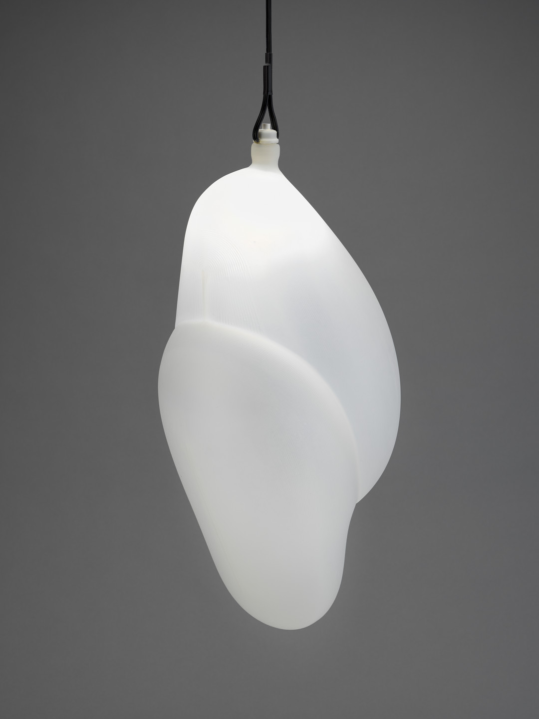 A white, smooth, and curvilinear pendant light hangs from a black cord against a gray background. The light has an organic shape with rounded, flowing contours. This is an AI generated description. To give feedback, email ai@moma.org.