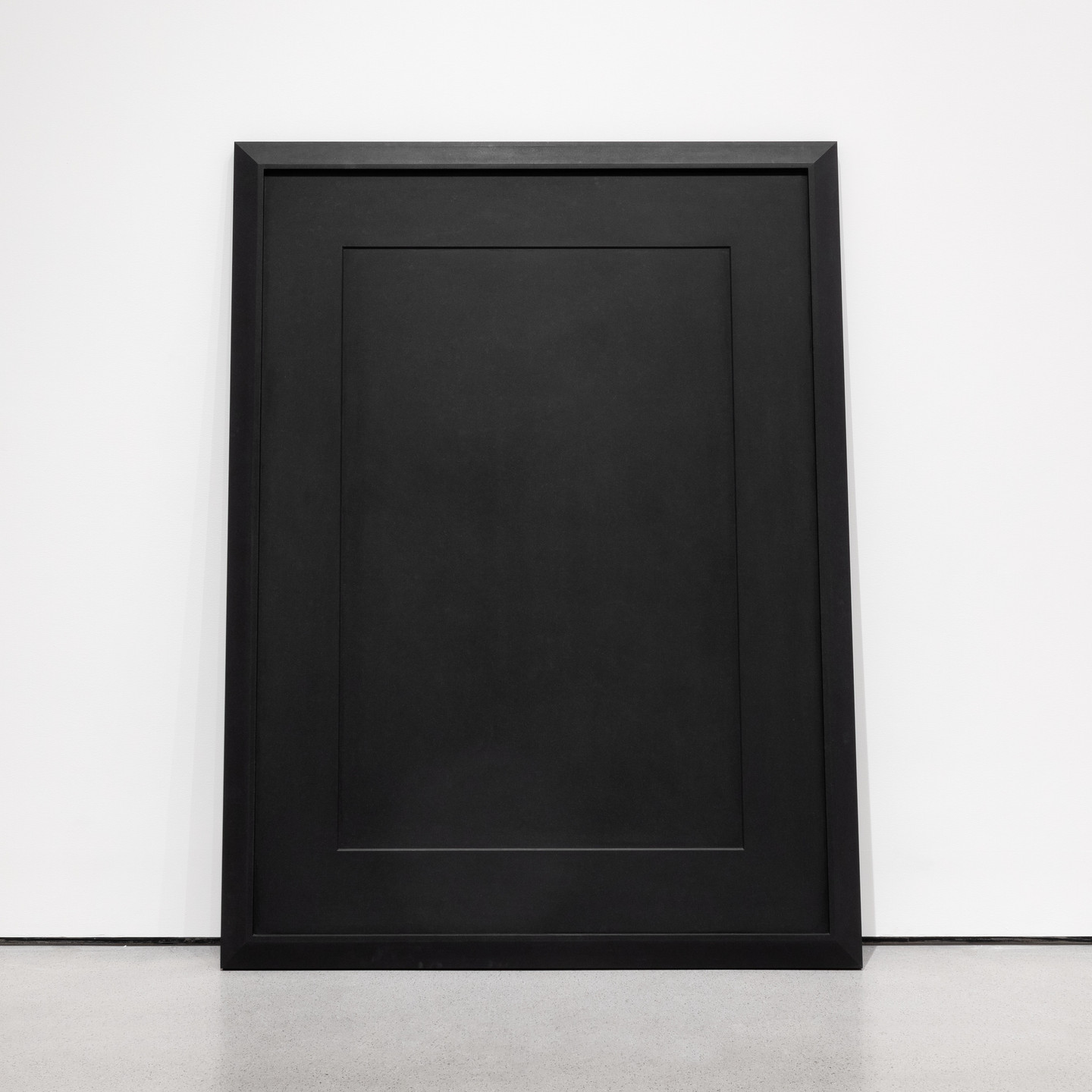 A large, black rectangular frame with a slightly raised inner border rests against a white wall. The floor beneath it is light gray and smooth. This is an AI generated description. To give feedback, email ai@moma.org.