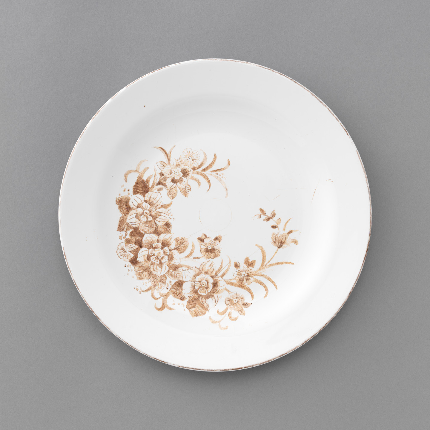 A white plate with a brown floral pattern on one side. The background is grey. This is an AI generated description. To give feedback, email ai@moma.org.