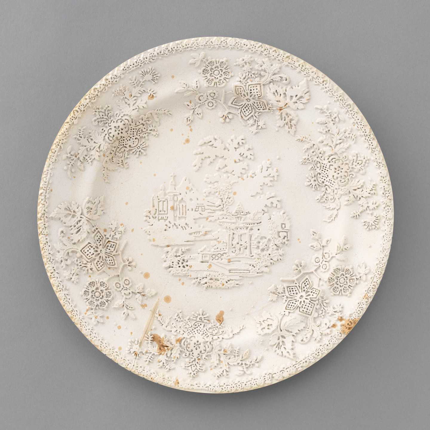 A circular, off-white plate with detailed floral patterns and a central scene depicting a village with trees and buildings. The plate has small brown spots scattered across its surface. This is an AI generated description. To give feedback, email ai@moma.org.